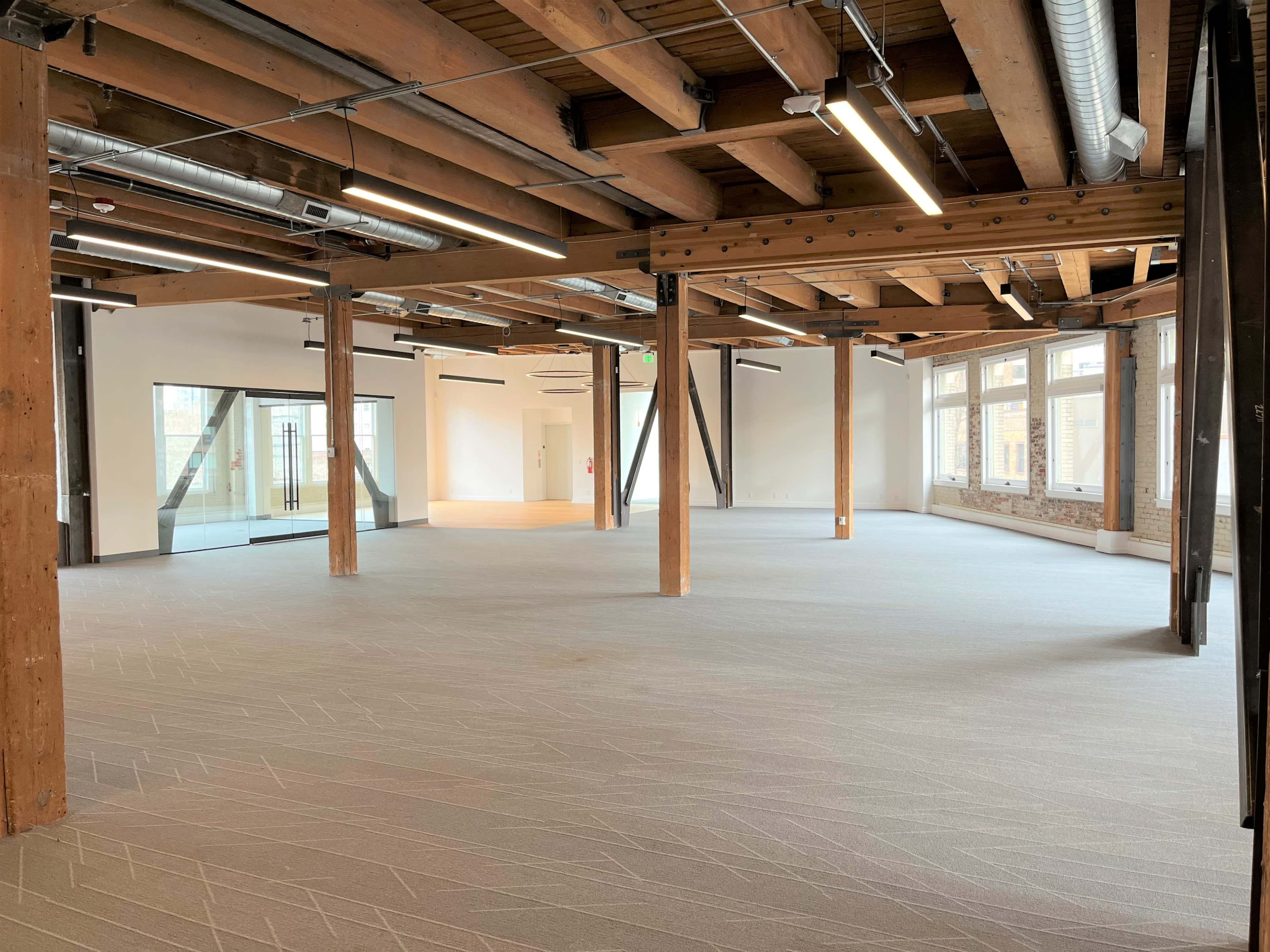 The image shows an empty office space with wooden beams, large windows, and light-colored carpet.