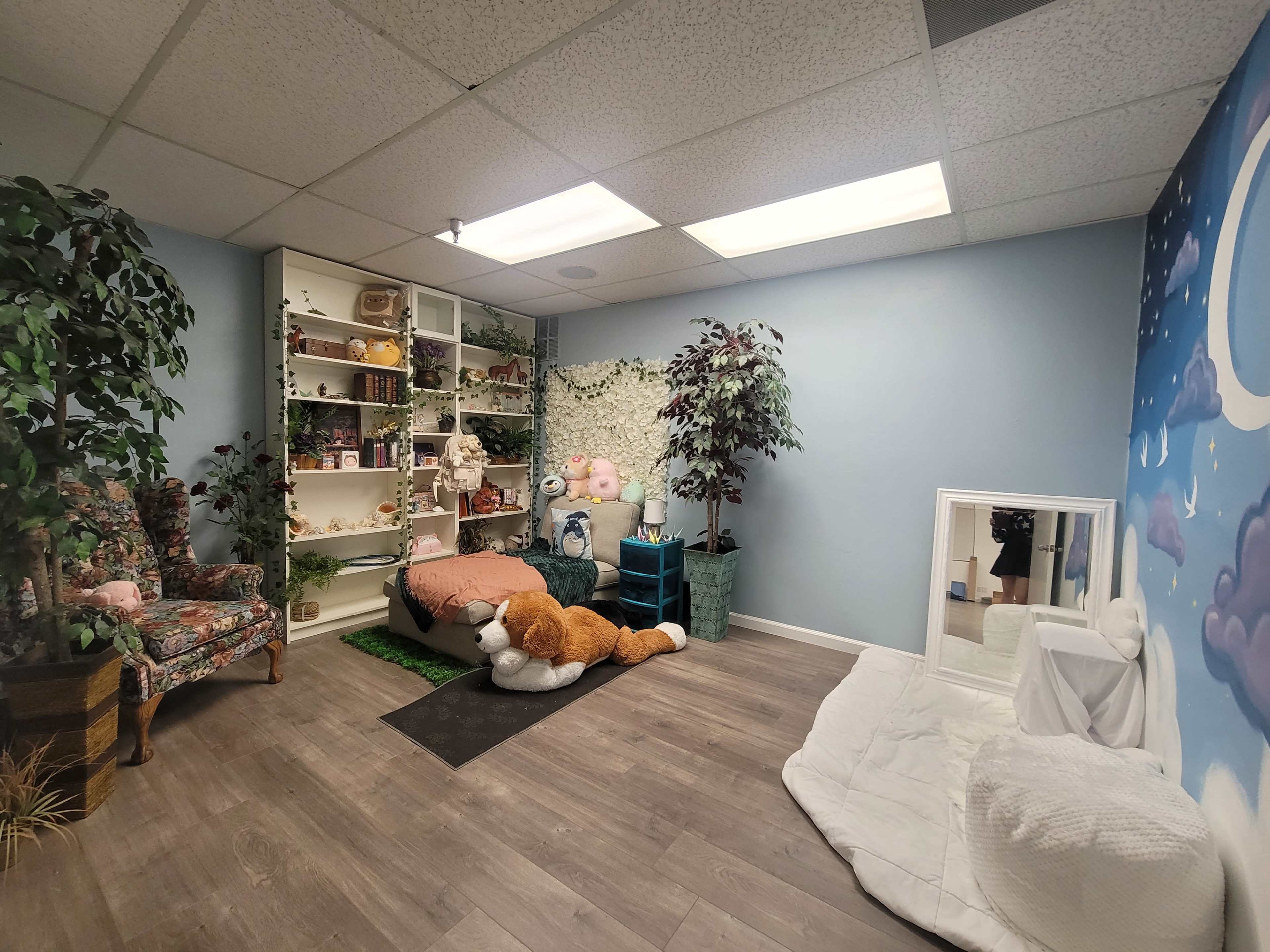 The image shows a cozy room with a colorful mural, a shelf filled with various items, a plush chair, a large stuffed animal, and a mirror on the floor near a white blanket.