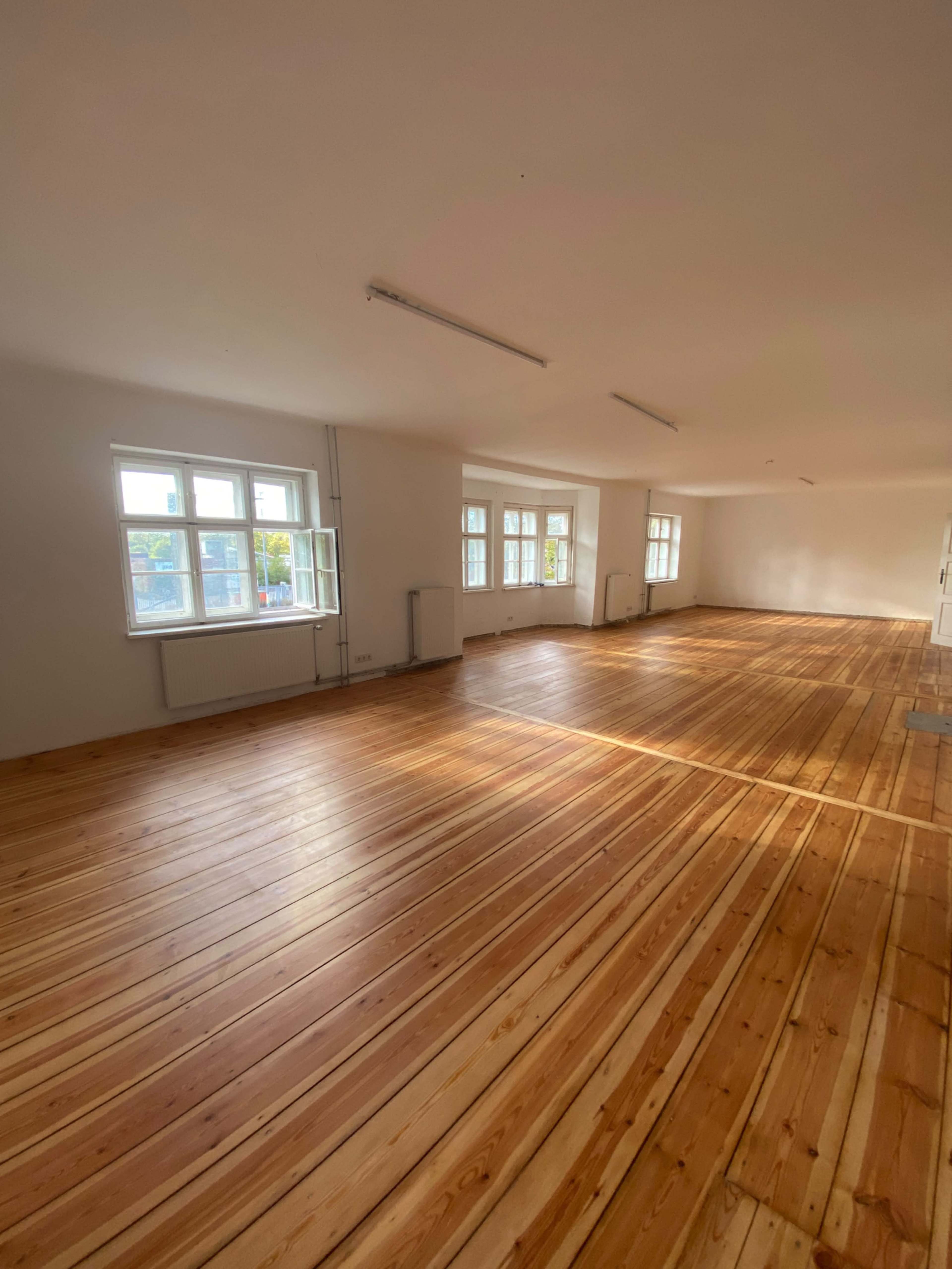 The image shows a spacious, empty room with wooden flooring and several large windows allowing natural light to enter.