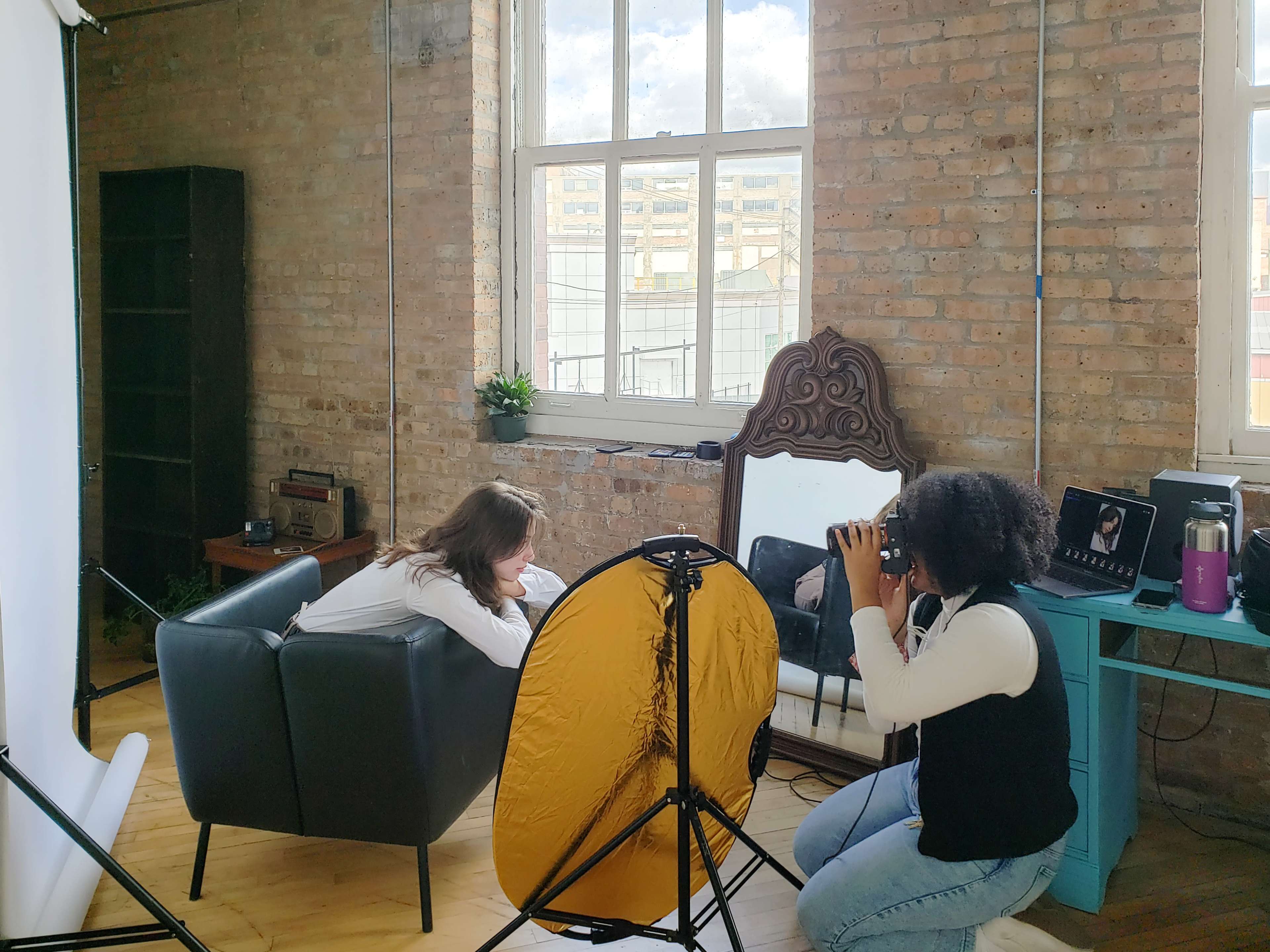 A photographer captures an image of a model sitting on a sofa in a studio setting with large windows and a backdrop.