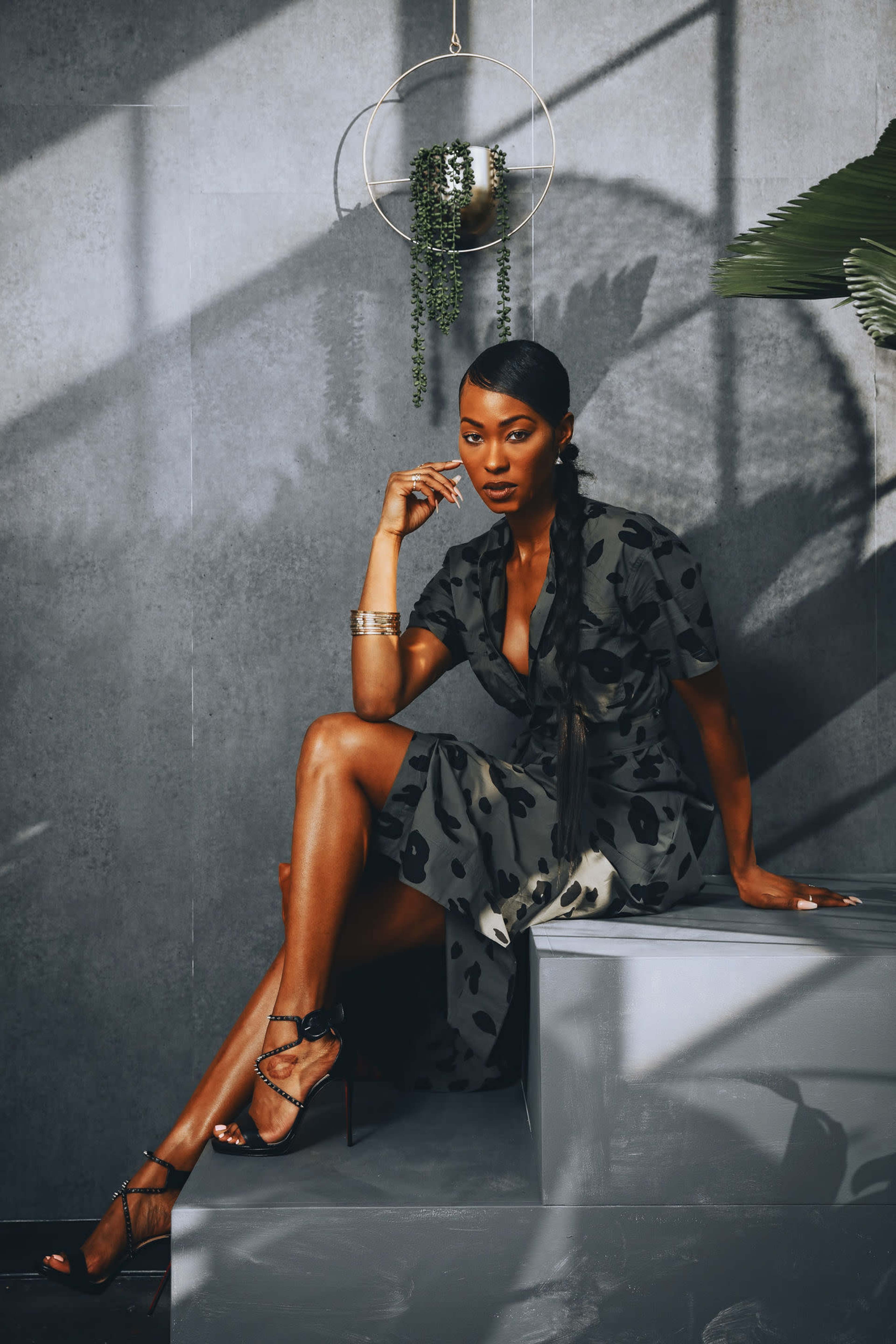 A woman sits on a gray step in a patterned dress with a plant and a hanging planter in the background.