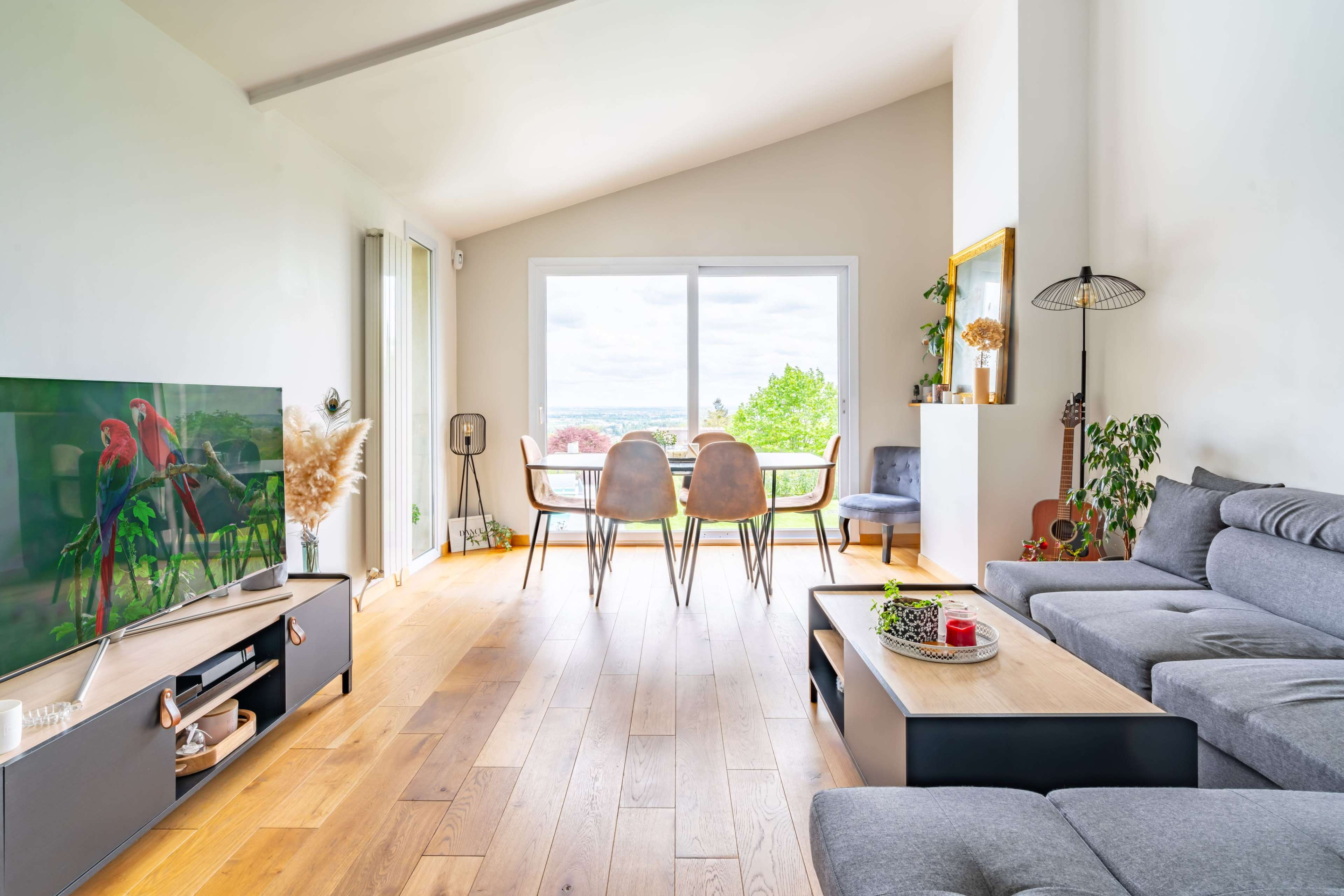 A modern living room featuring a gray sofa, a wooden floor, a large window with a view, and a dining table set for four.