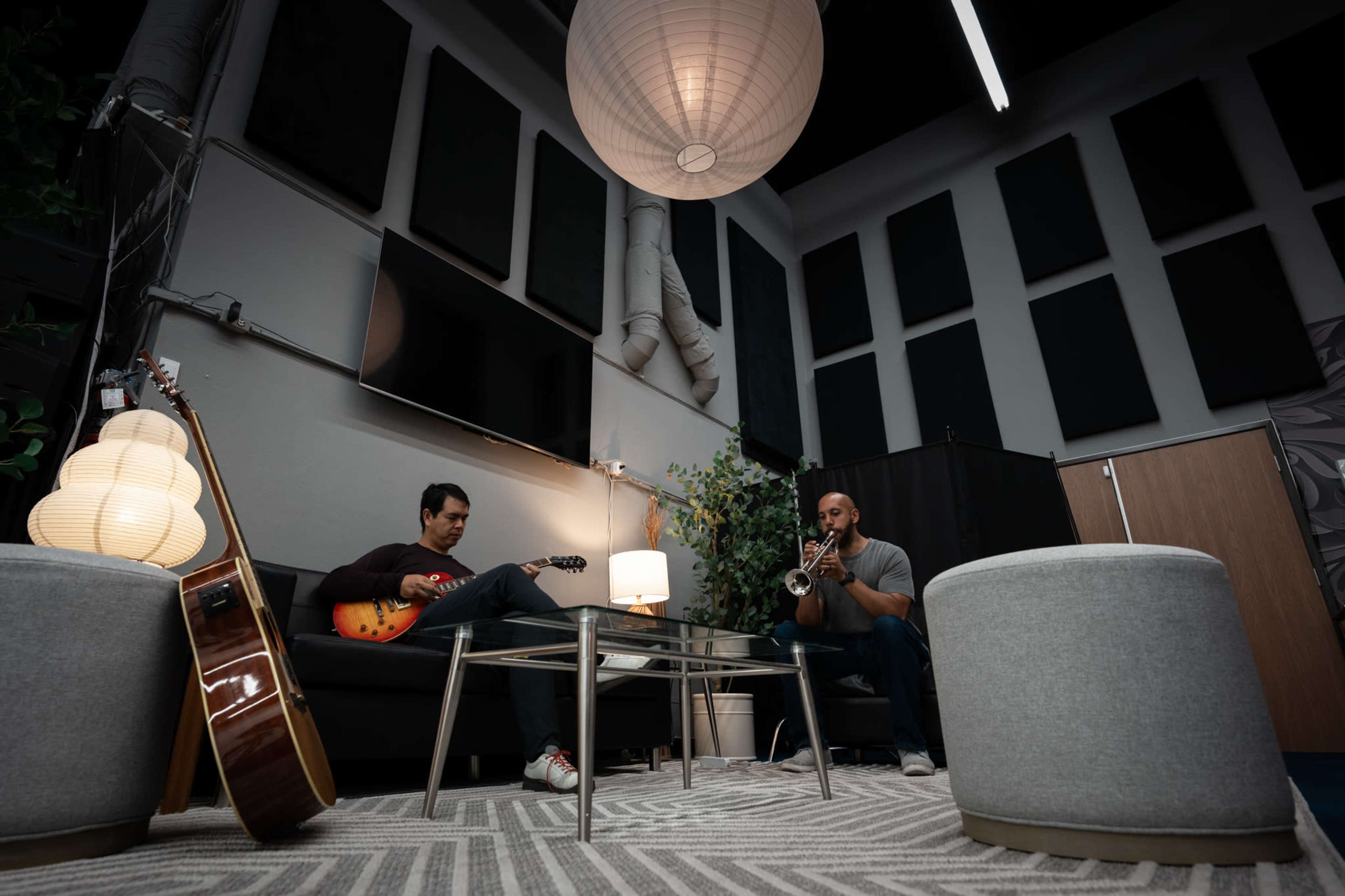 Two musicians are seated in a modern lounge, one playing an electric guitar and the other playing a trumpet, surrounded by ambient lighting and acoustic panels.