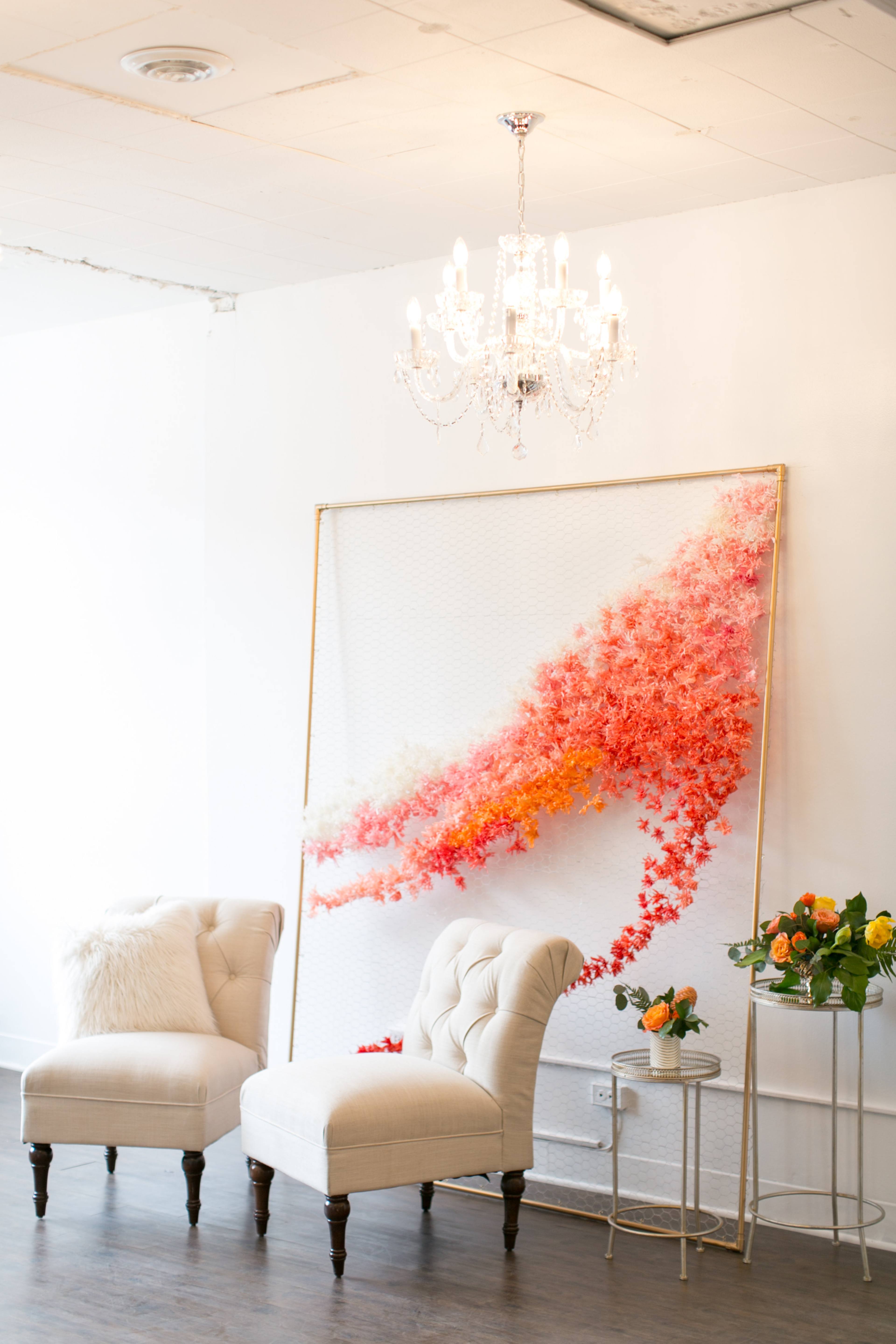 The scene features two upholstered chairs beside a floral wall art piece and a side table with a bouquet, all set under a chandelier in a bright room.