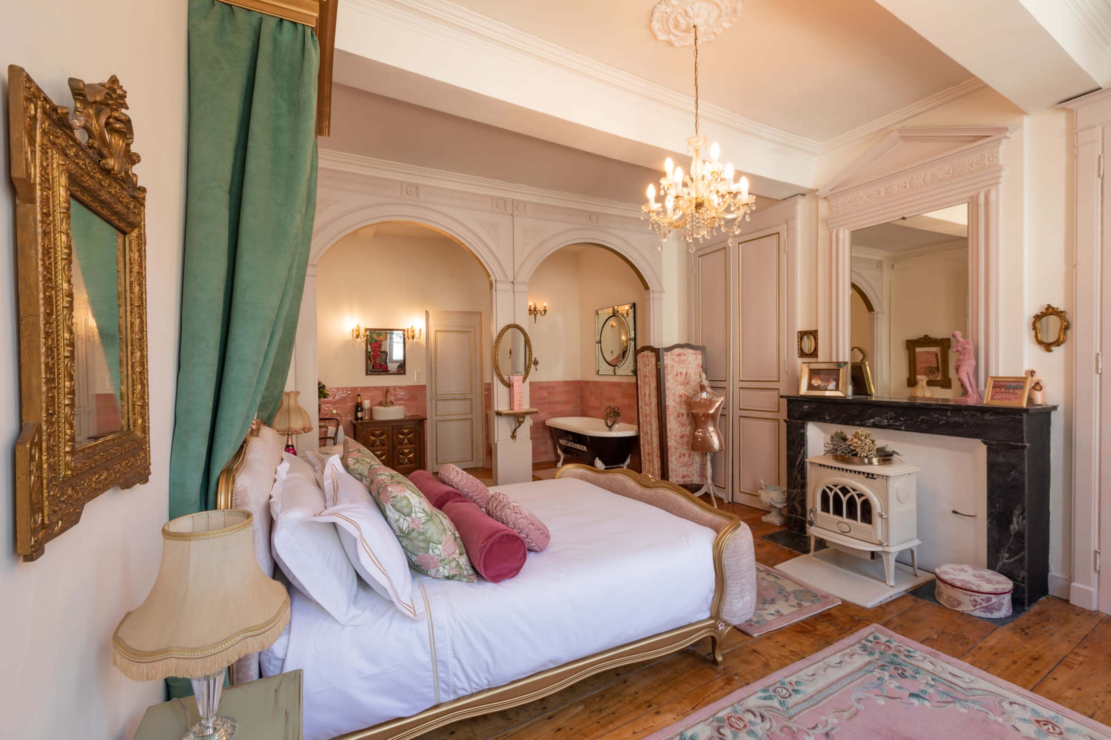 The image shows a elegantly decorated bedroom featuring a large bed, vintage-style mirrors, a chandelier, and a decorative fireplace.