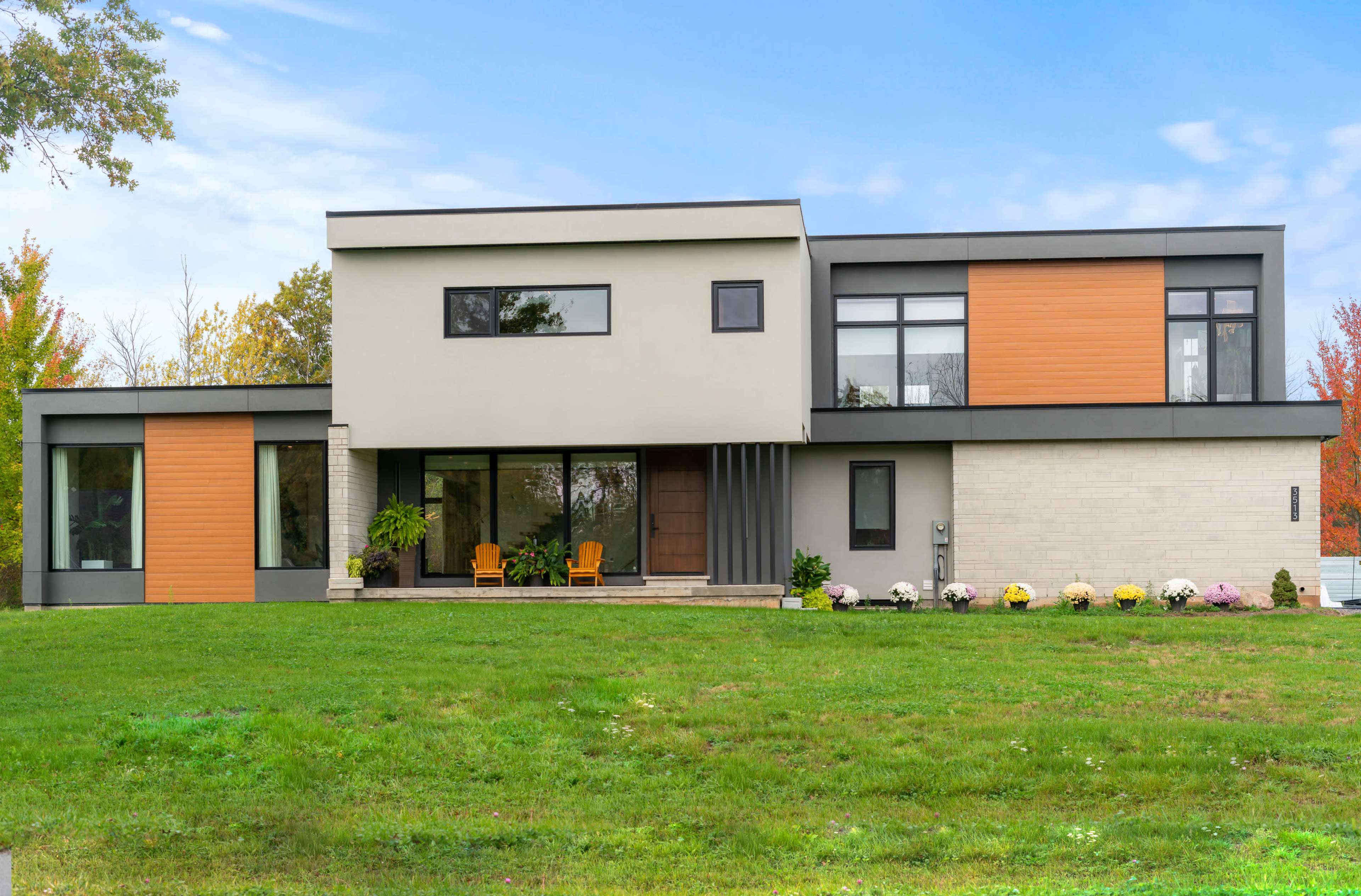 A modern two-story house features a mix of gray and wooden panel exteriors, large windows, and a front porch with two chairs.