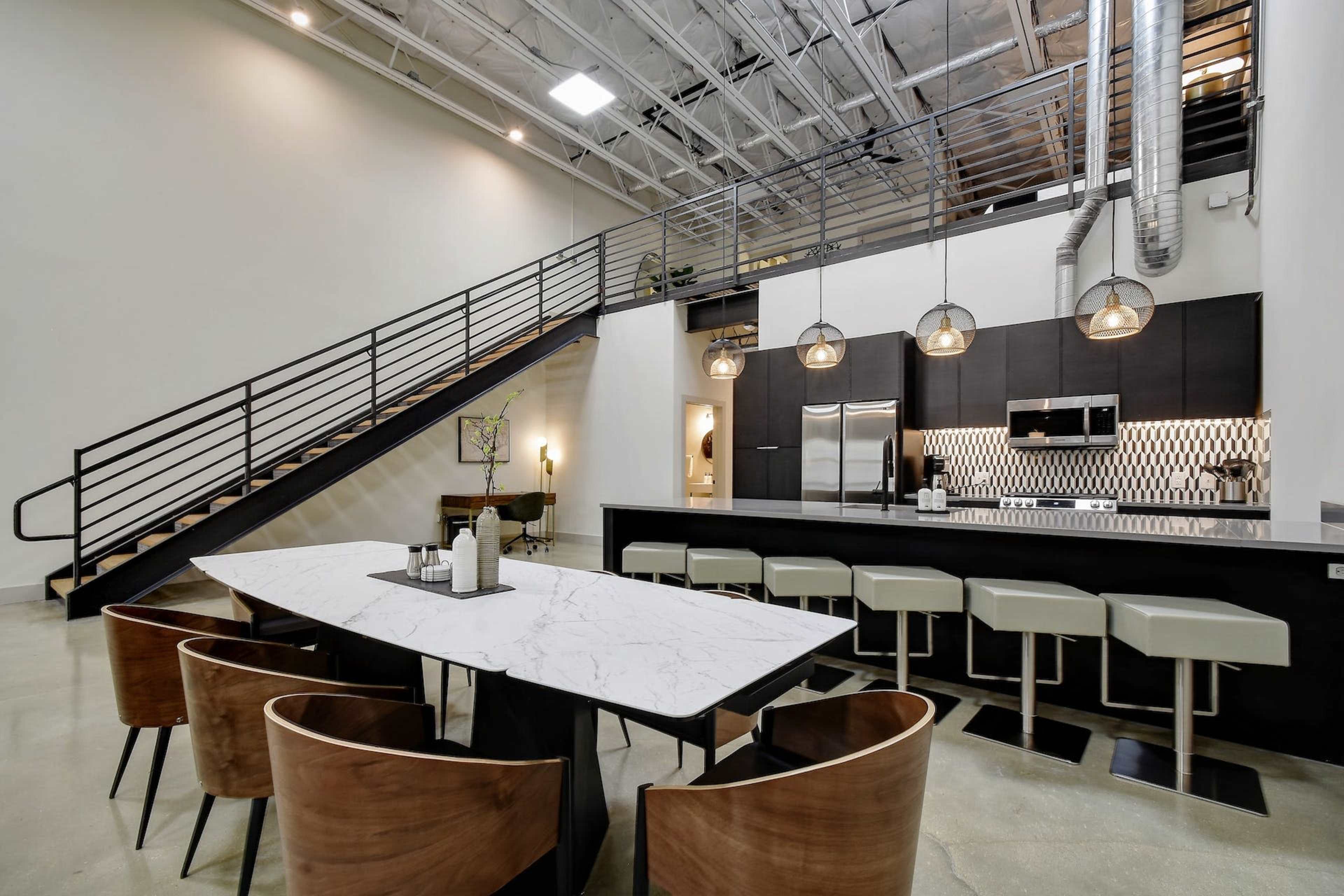 A modern open-concept kitchen features a marble dining table surrounded by wooden chairs, with a staircase in the background and pendant lights above the counter.