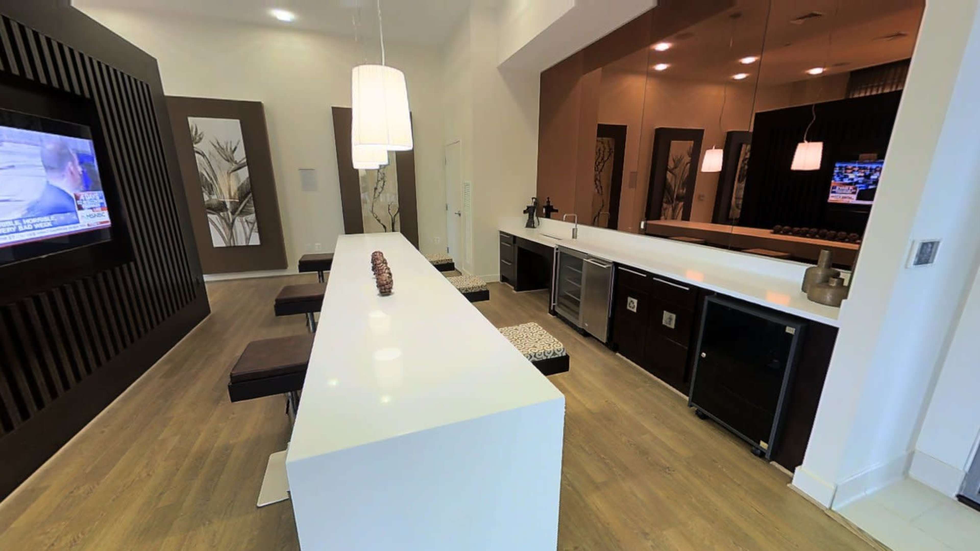 A modern kitchen area with a long white countertop, dark cabinets, and a television on the wall.