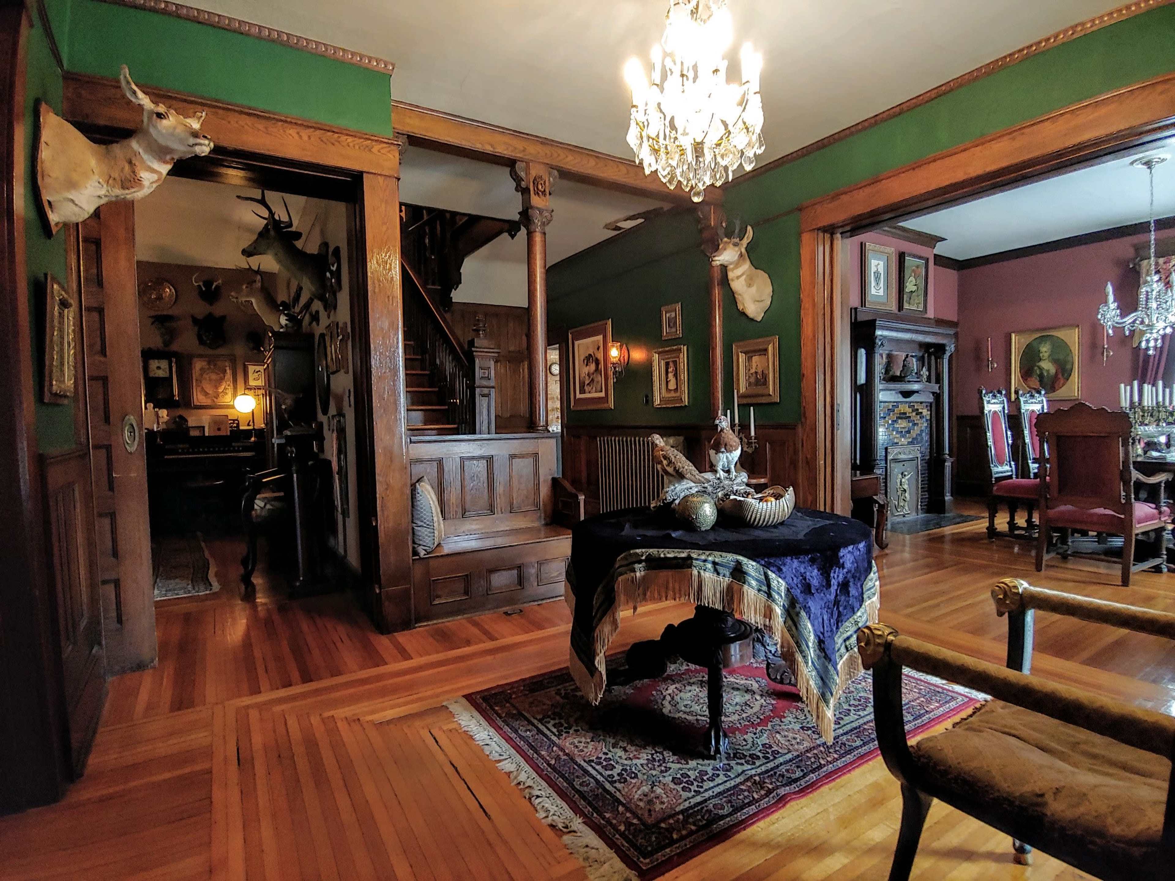 The image depicts a vintage interior featuring wooden walls, a staircase, and decorative elements including mounted animal heads and ornate furniture.