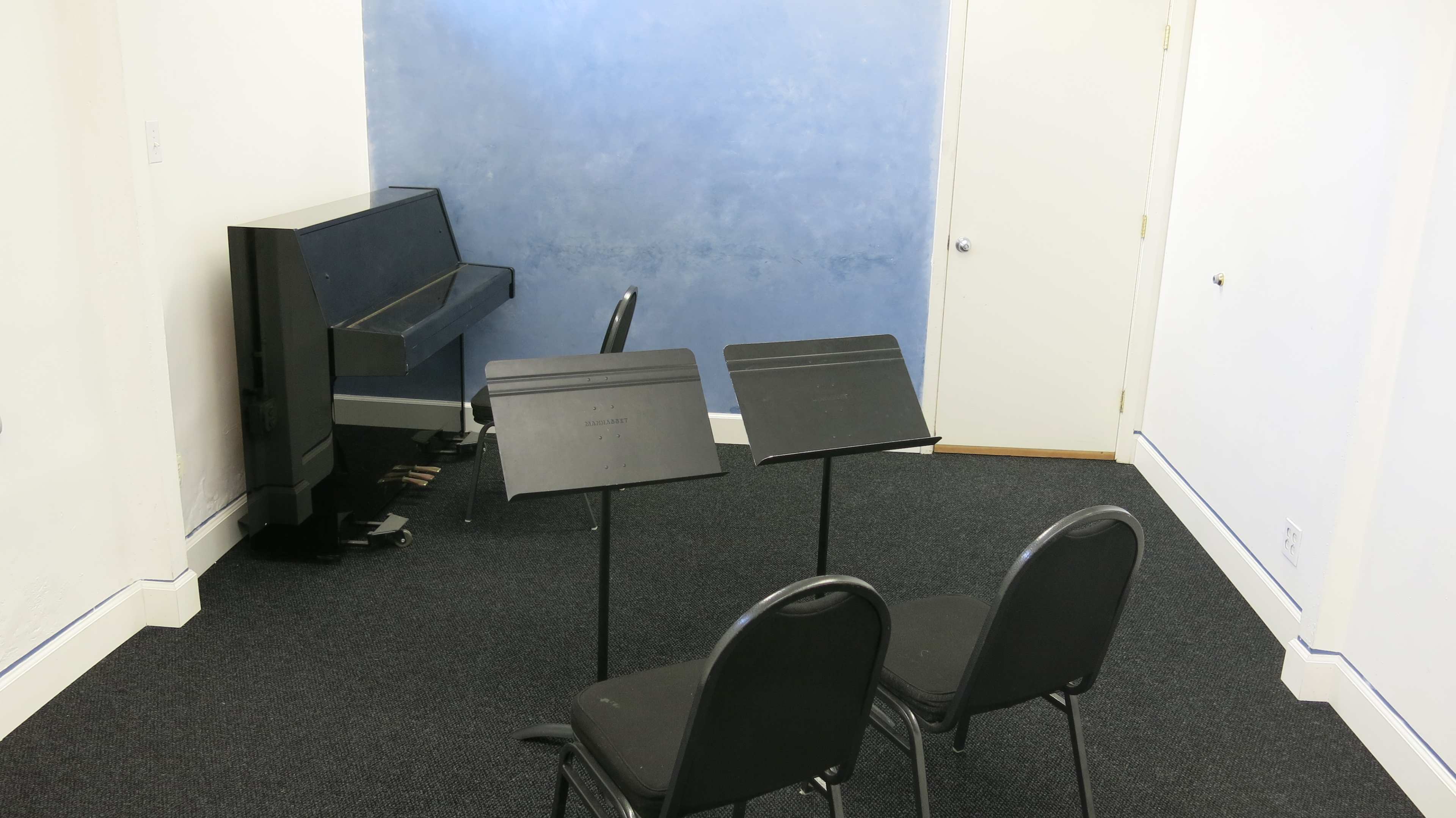 A small room with a black piano, three music stands, and a chair against a blue wall.
