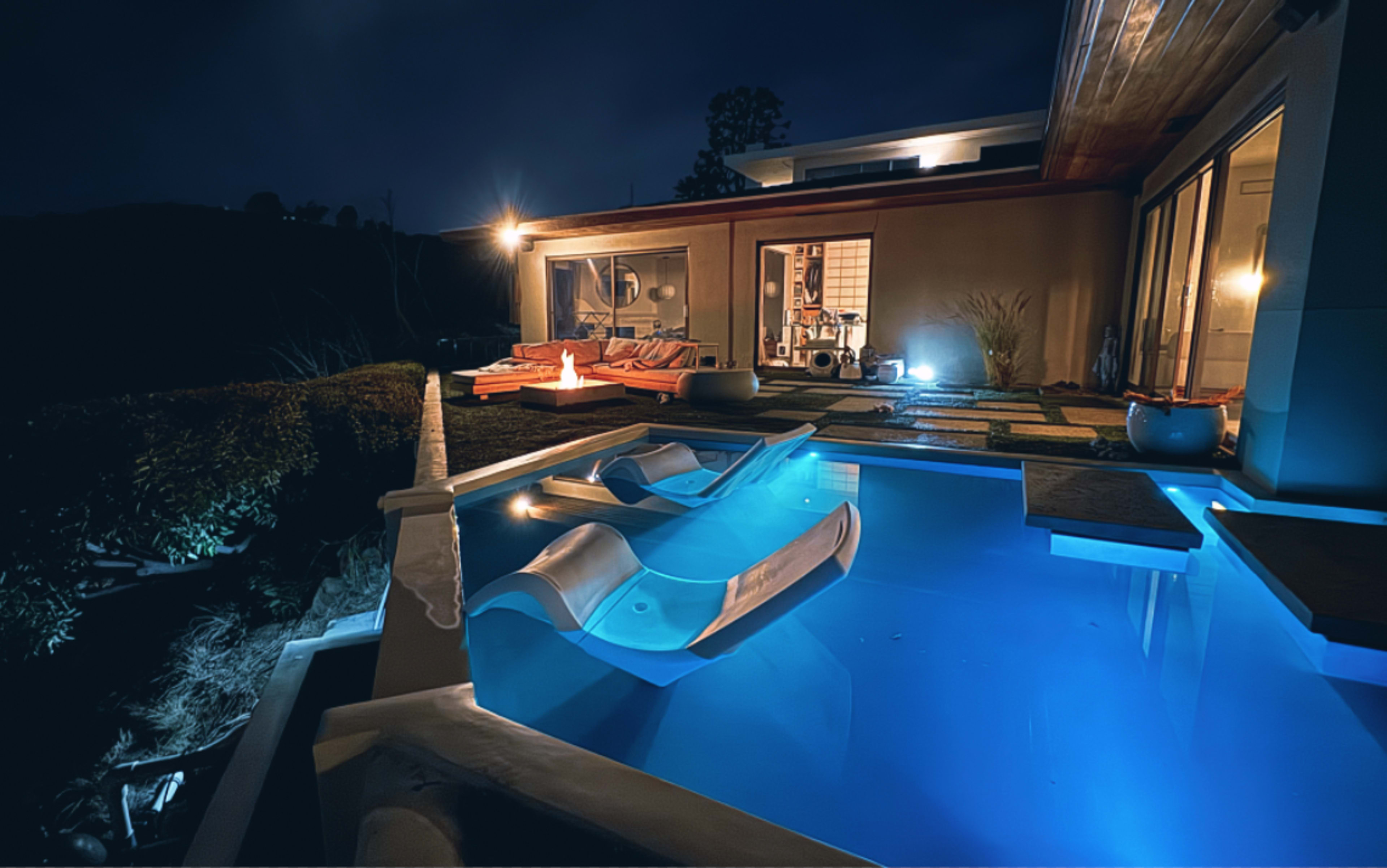 The image shows a modern villa at night with a well-lit swimming pool featuring two water slides and a cozy outdoor seating area.