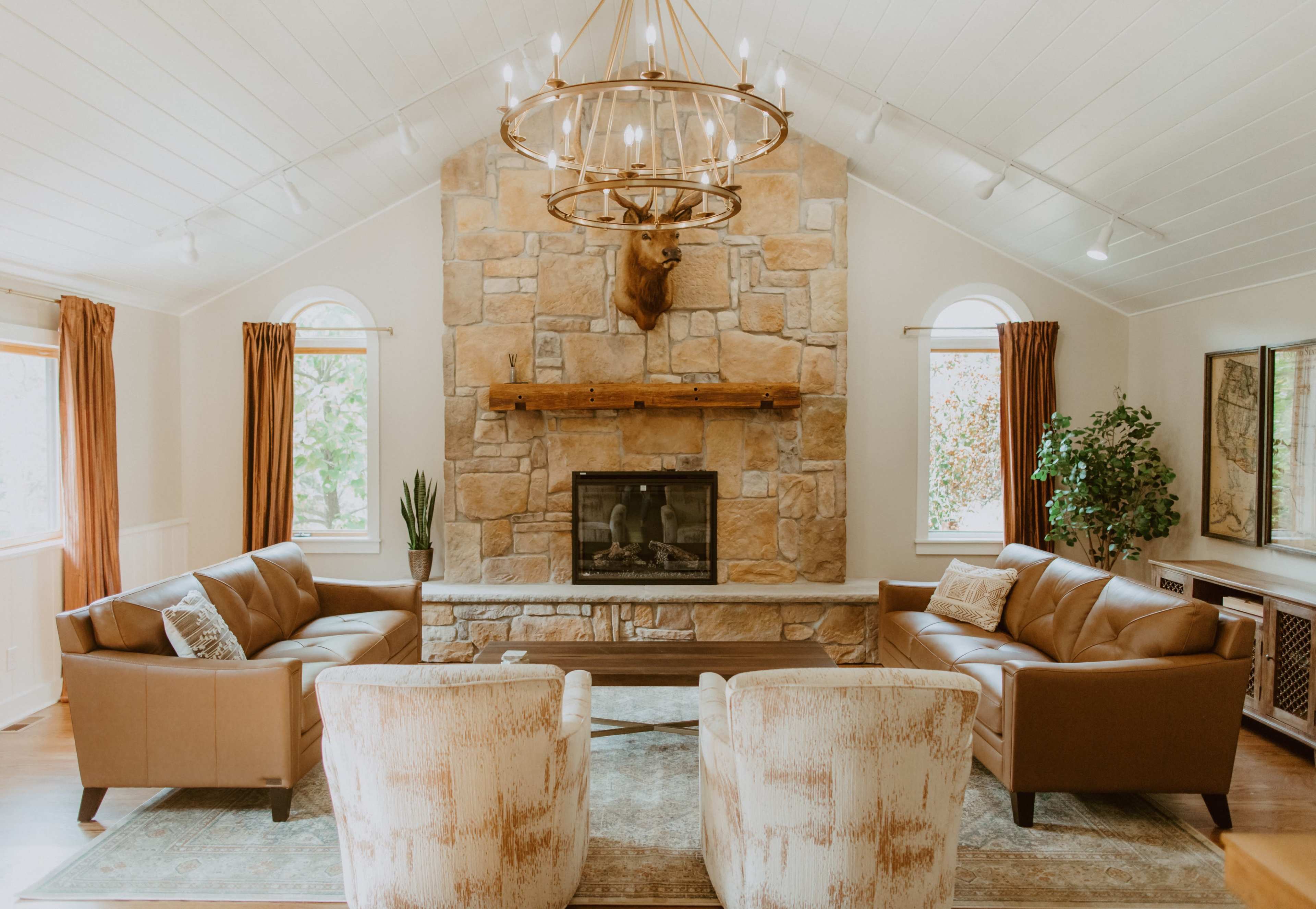 The living room features two leather sofas facing a stone fireplace with a mounted deer head above it, flanked by windows and decorative plants.