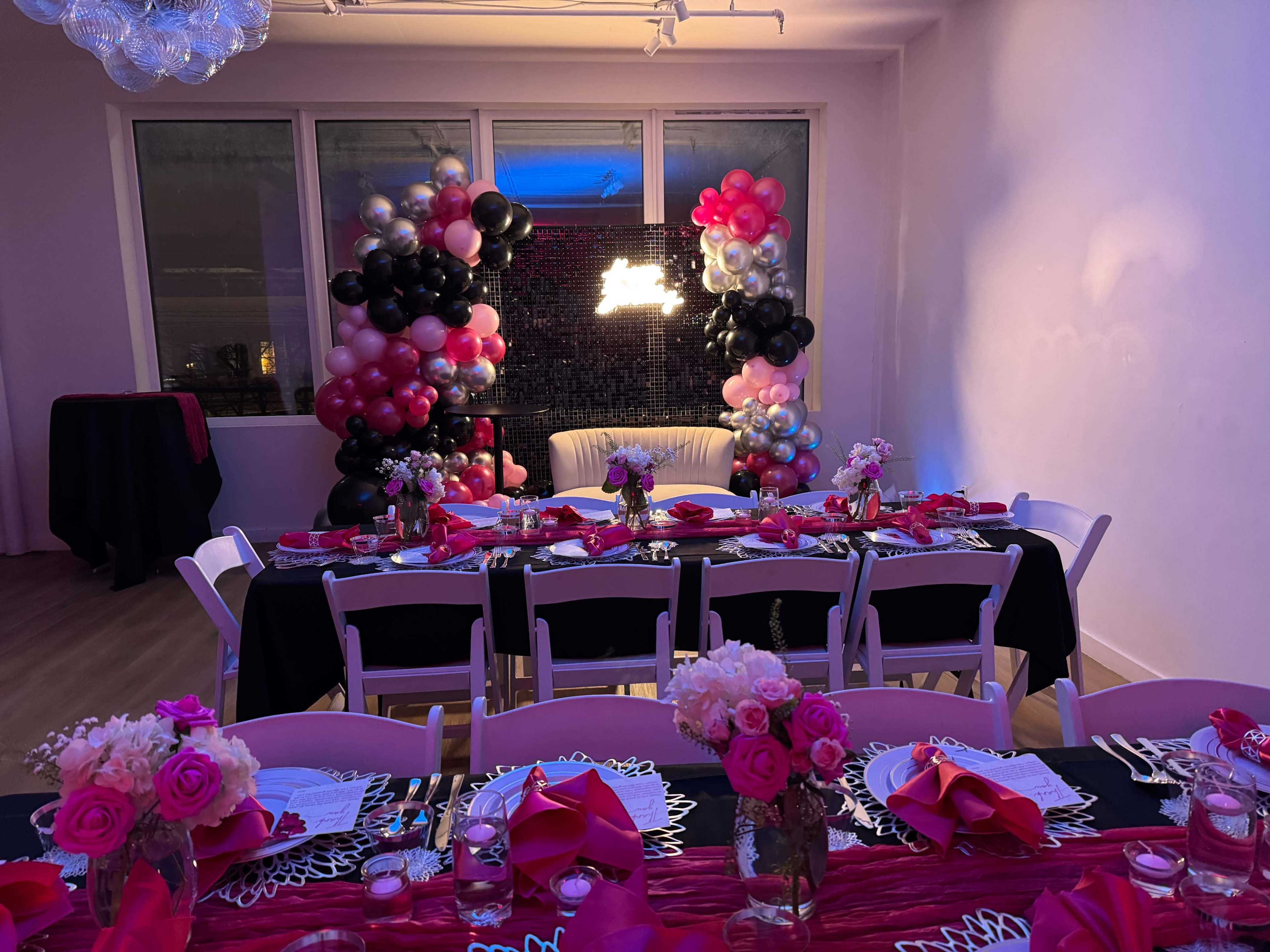The image shows a decorated event space with a long table set for a celebration, surrounded by colorful balloons and floral centerpieces.