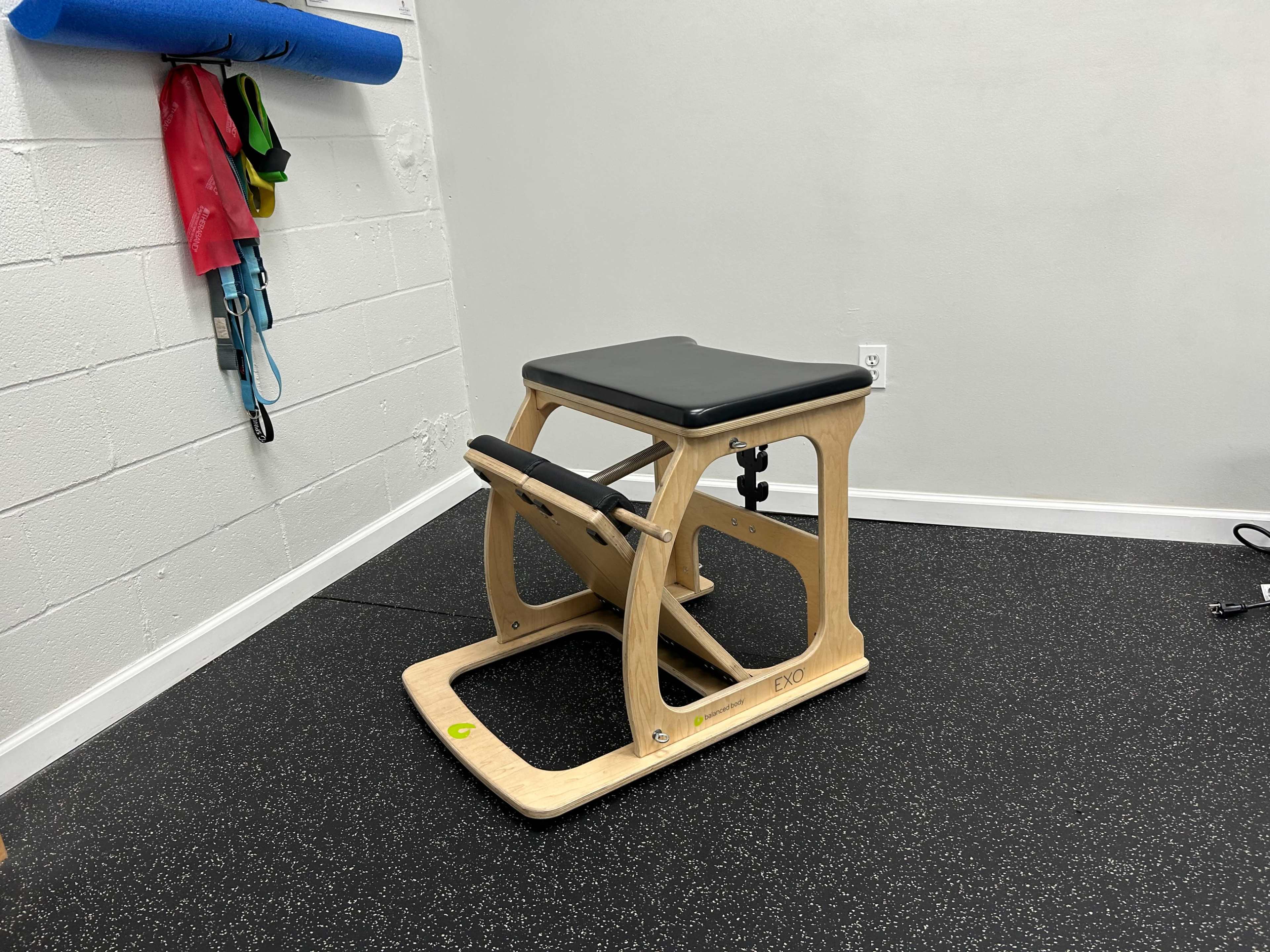 A wooden exercise equipment piece with a padded top and a footrest, placed in a room with dark rubber flooring and a white wall.