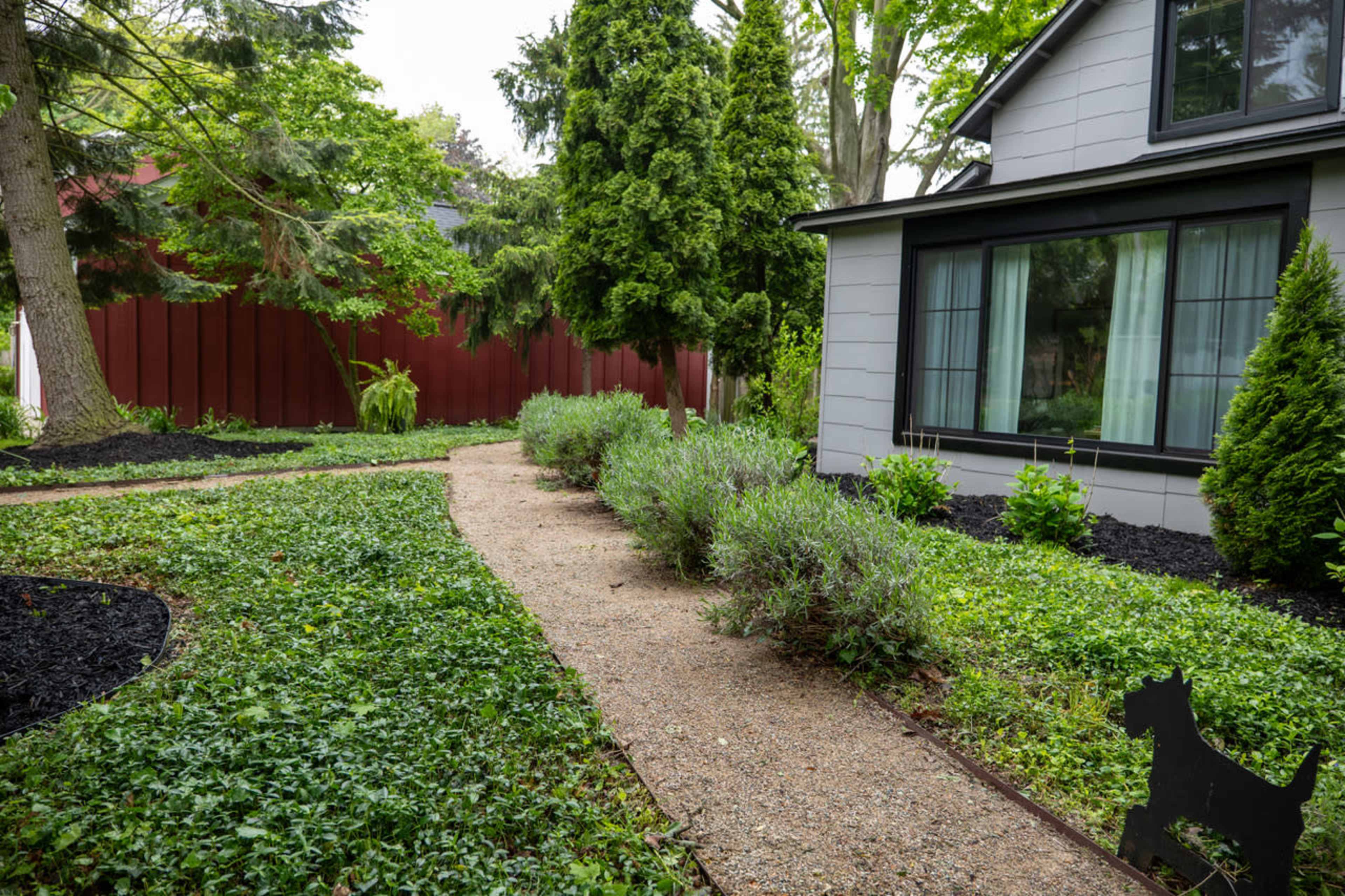 A gravel pathway winds through a landscaped garden with green shrubs and trees alongside a house with large windows.