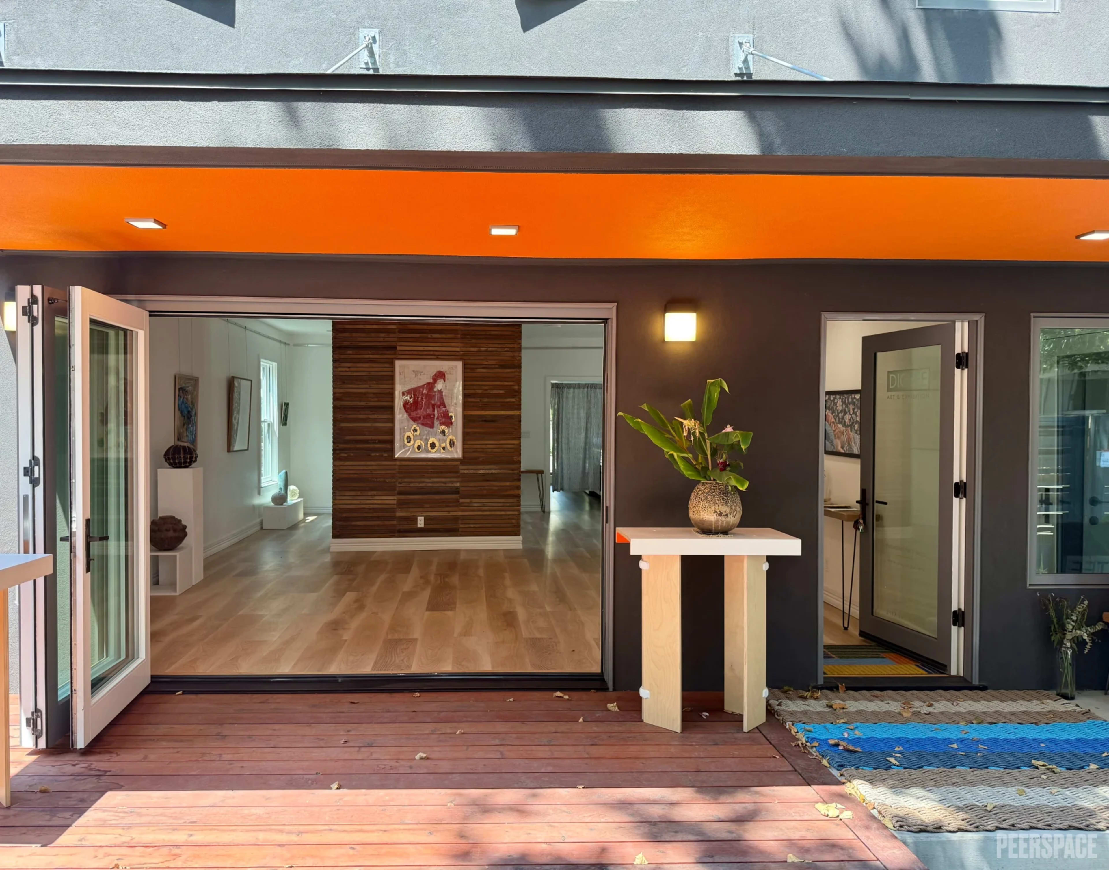 The image shows an open space with large glass doors leading to a wooden deck, framed by a modern interior featuring a mix of light and dark wood accents.