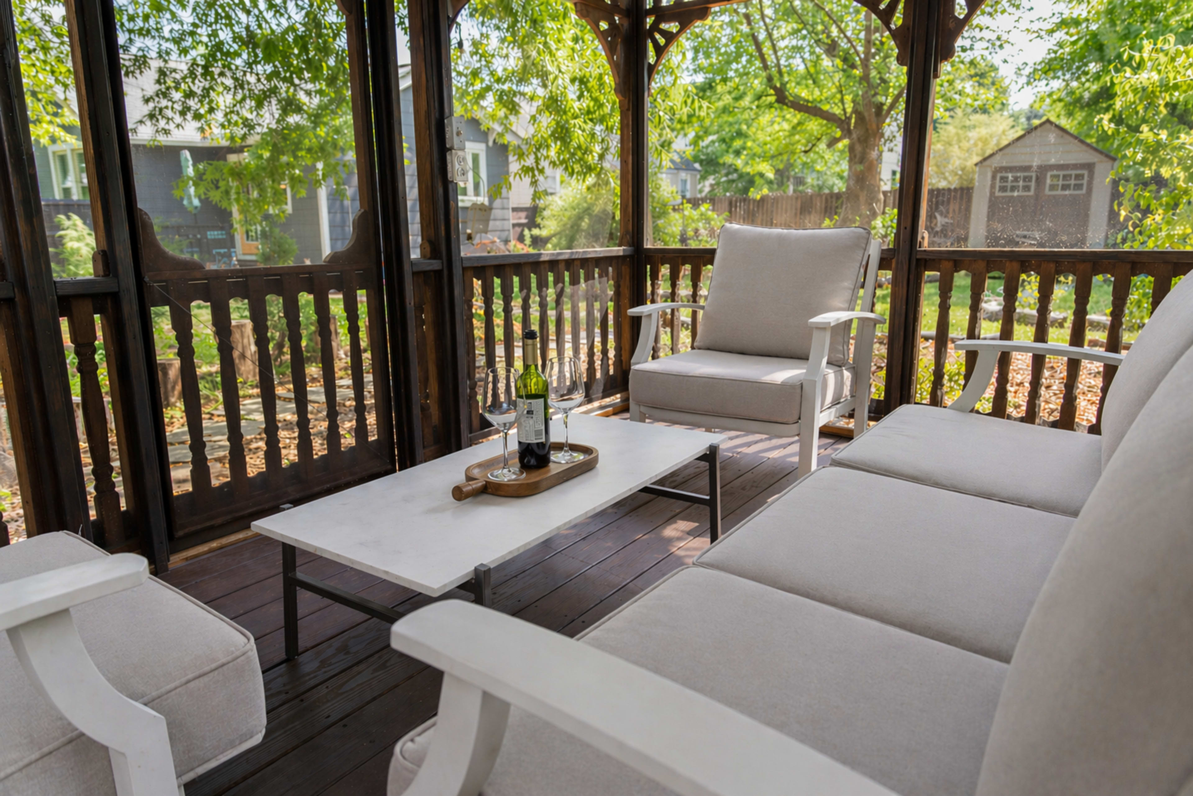 A cozy outdoor seating area features white cushioned chairs and a marble-top coffee table, surrounded by greenery.
