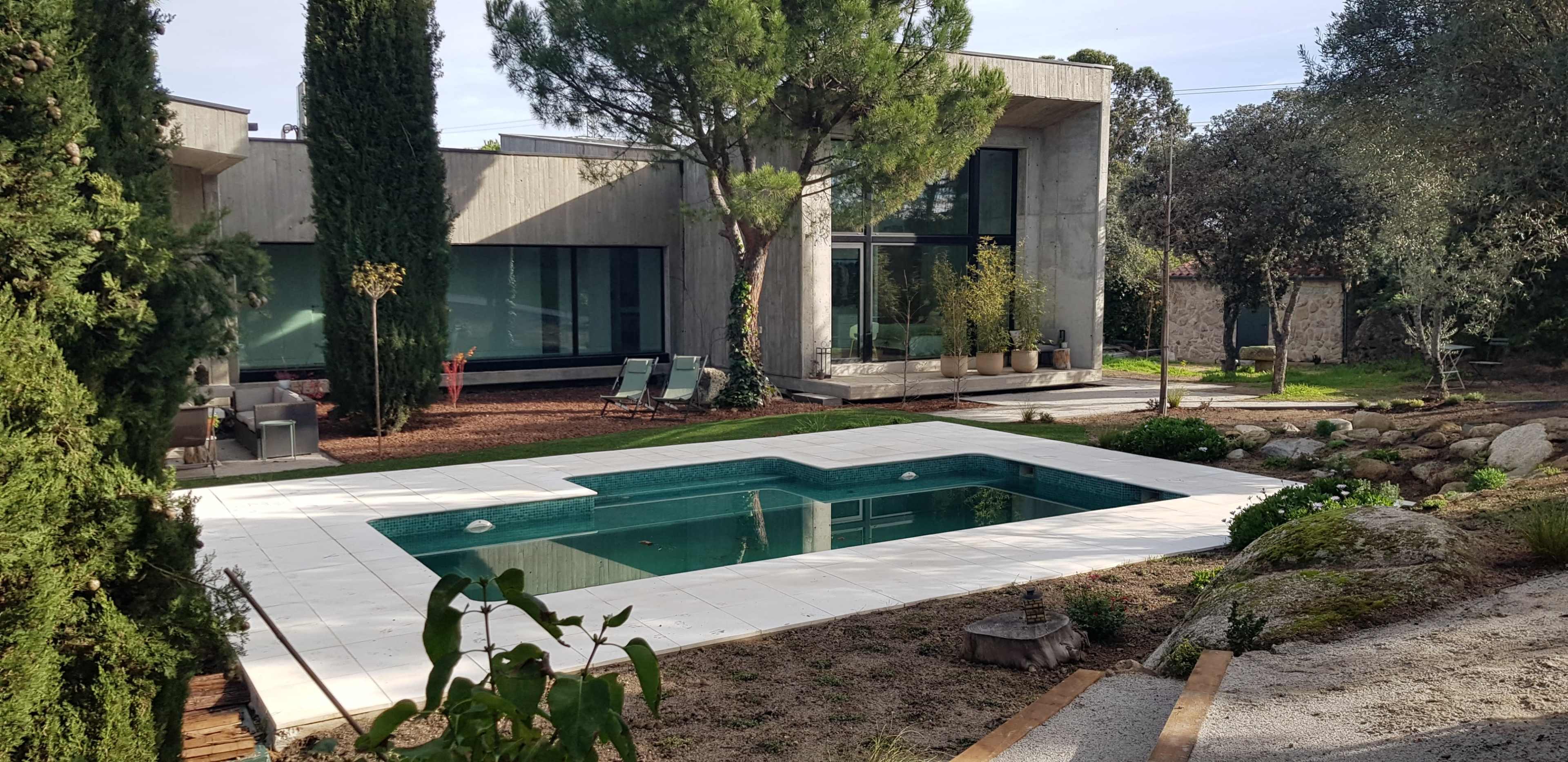 The image shows a modern house with large windows surrounded by greenery and a rectangular swimming pool in the foreground.