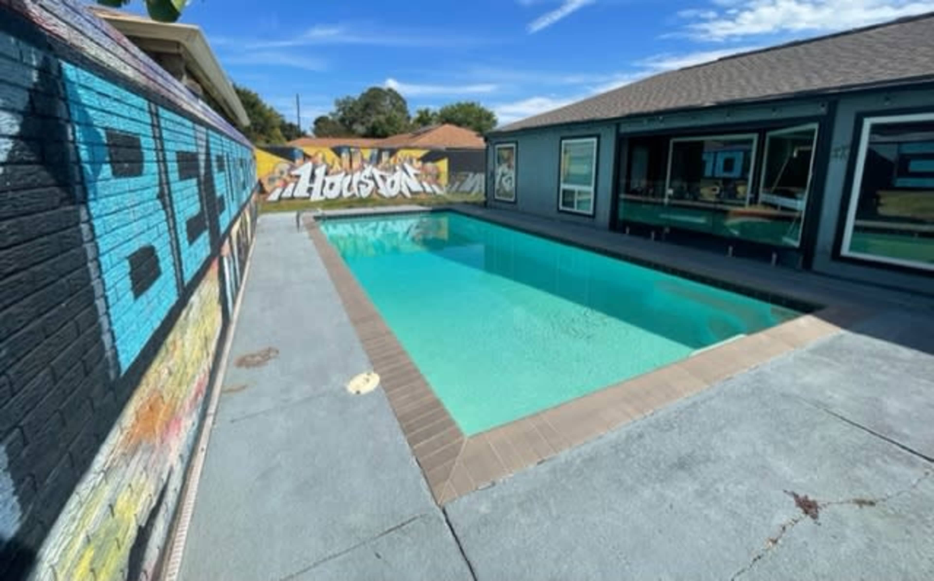 A clear swimming pool is surrounded by a concrete deck and a colorful mural on one side of the yard.