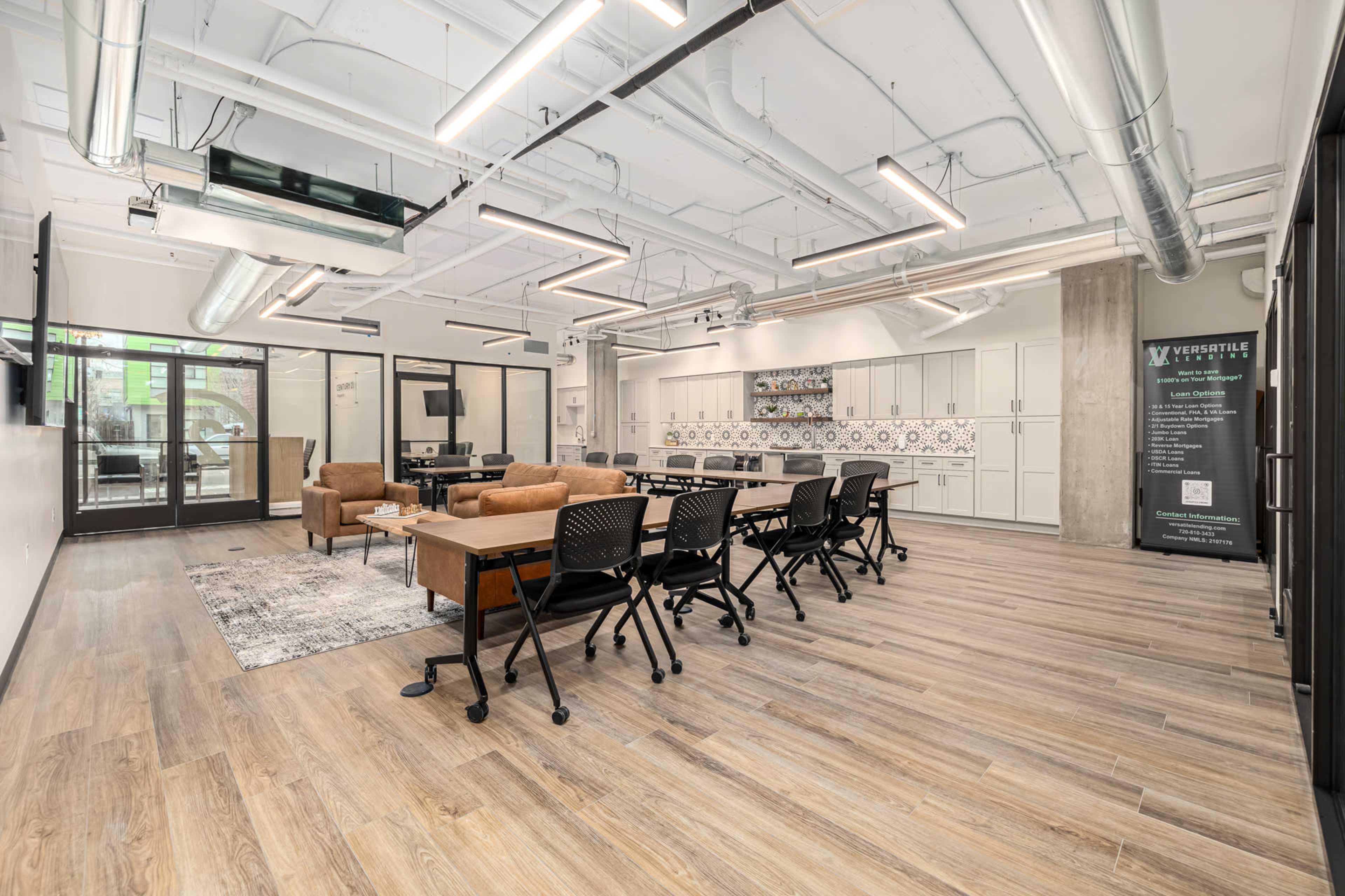 The image shows a modern office space with wooden flooring, multiple tables and chairs set up for meetings, and a lounge area with sofas.