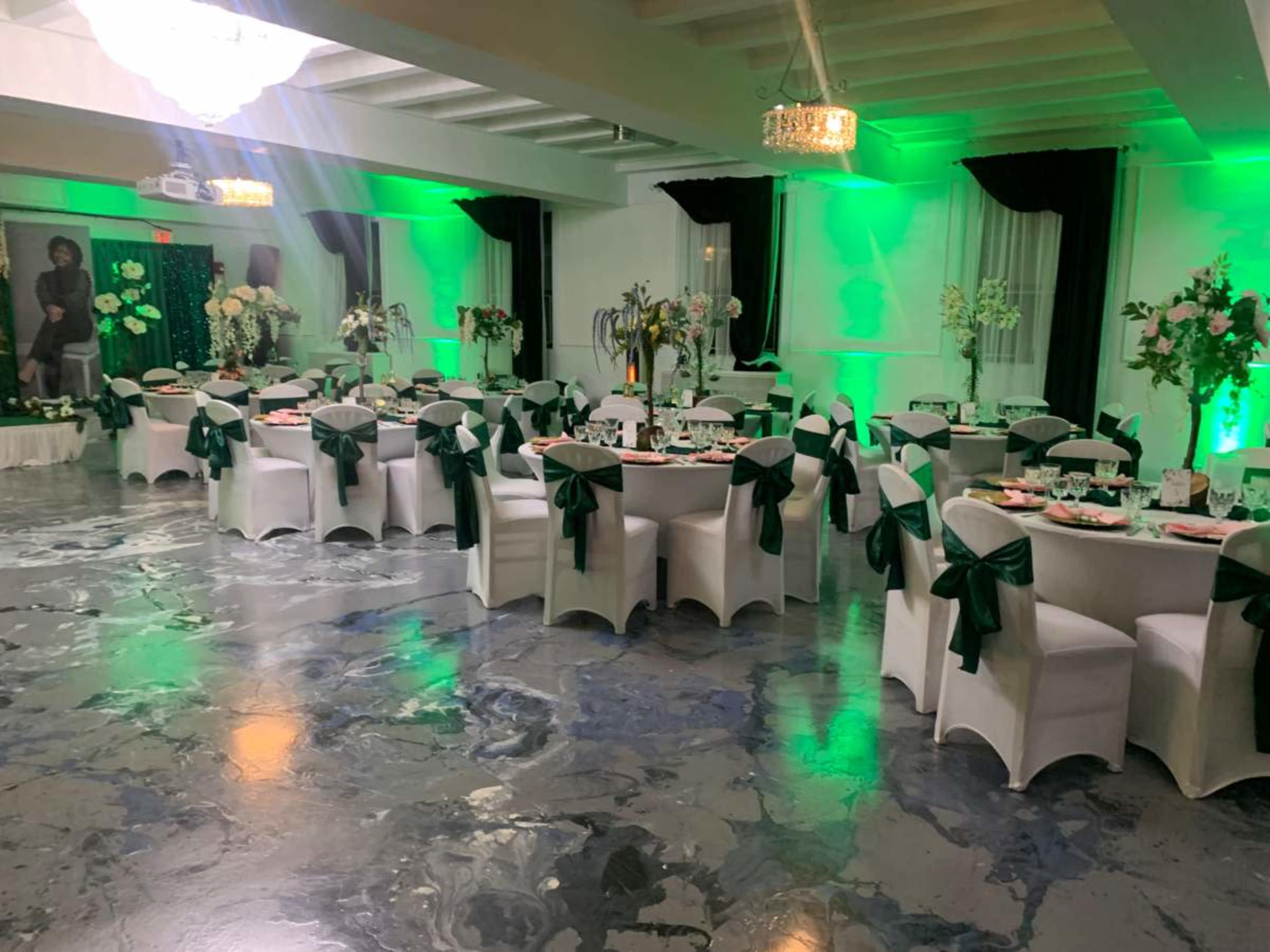 A banquet hall is set up with round tables dressed in white linens and green bows, surrounded by floral centerpieces and illuminated by green lighting.