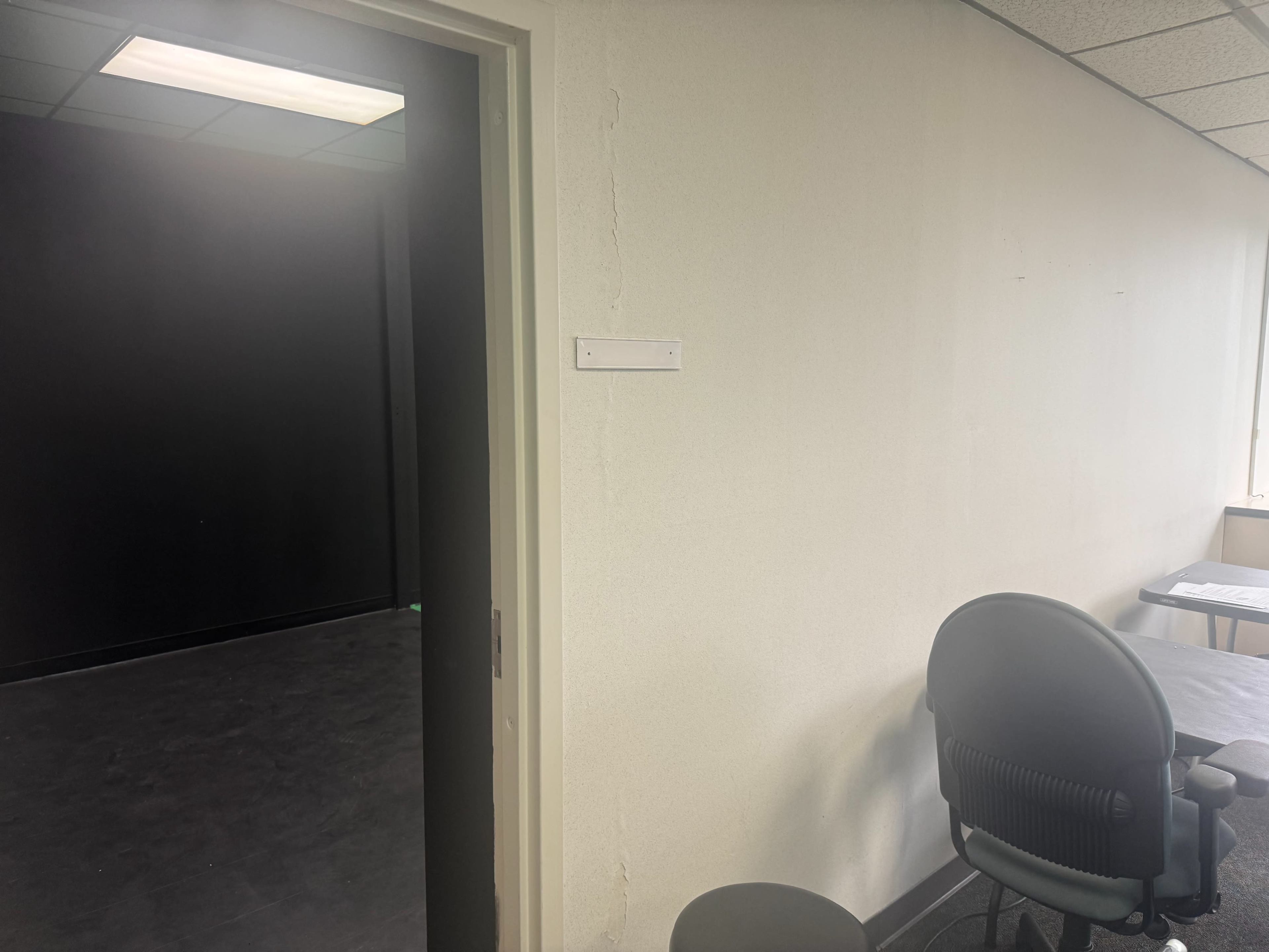 A doorway leads to a dark room visible from a brightly lit office space with a chair and table nearby.