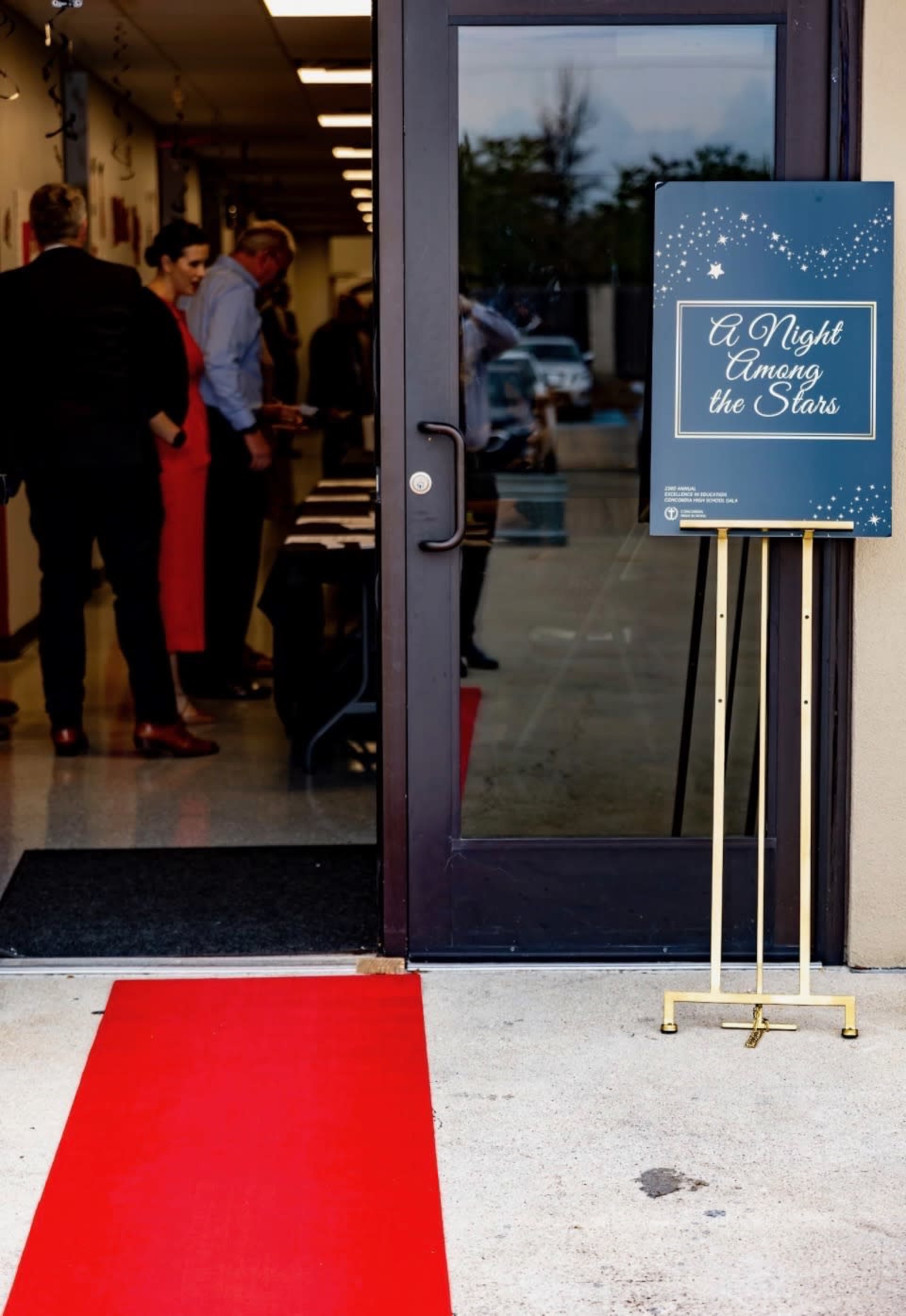 A red carpet leads to an entrance where a sign displays "A Night Among the Stars," while guests are engaged in conversation inside.