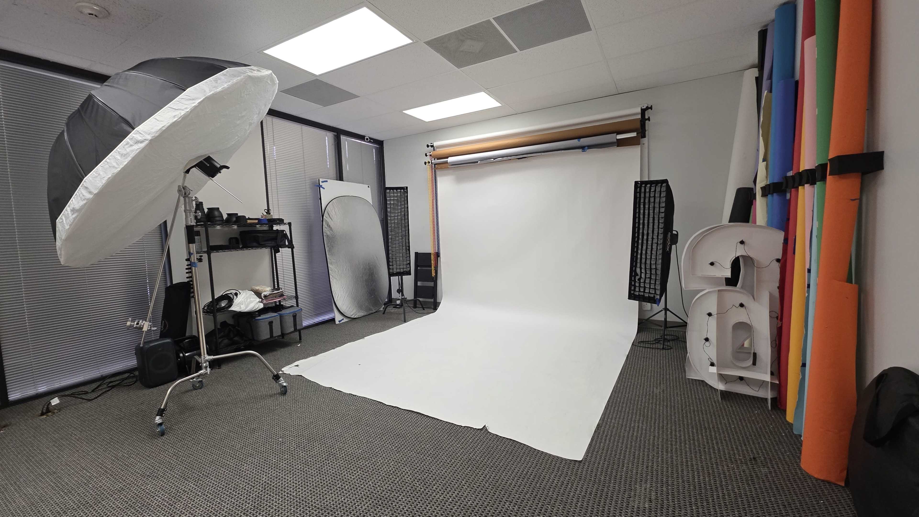 The image shows a photography studio equipped with a white backdrop, softbox lights, and various props against the walls.