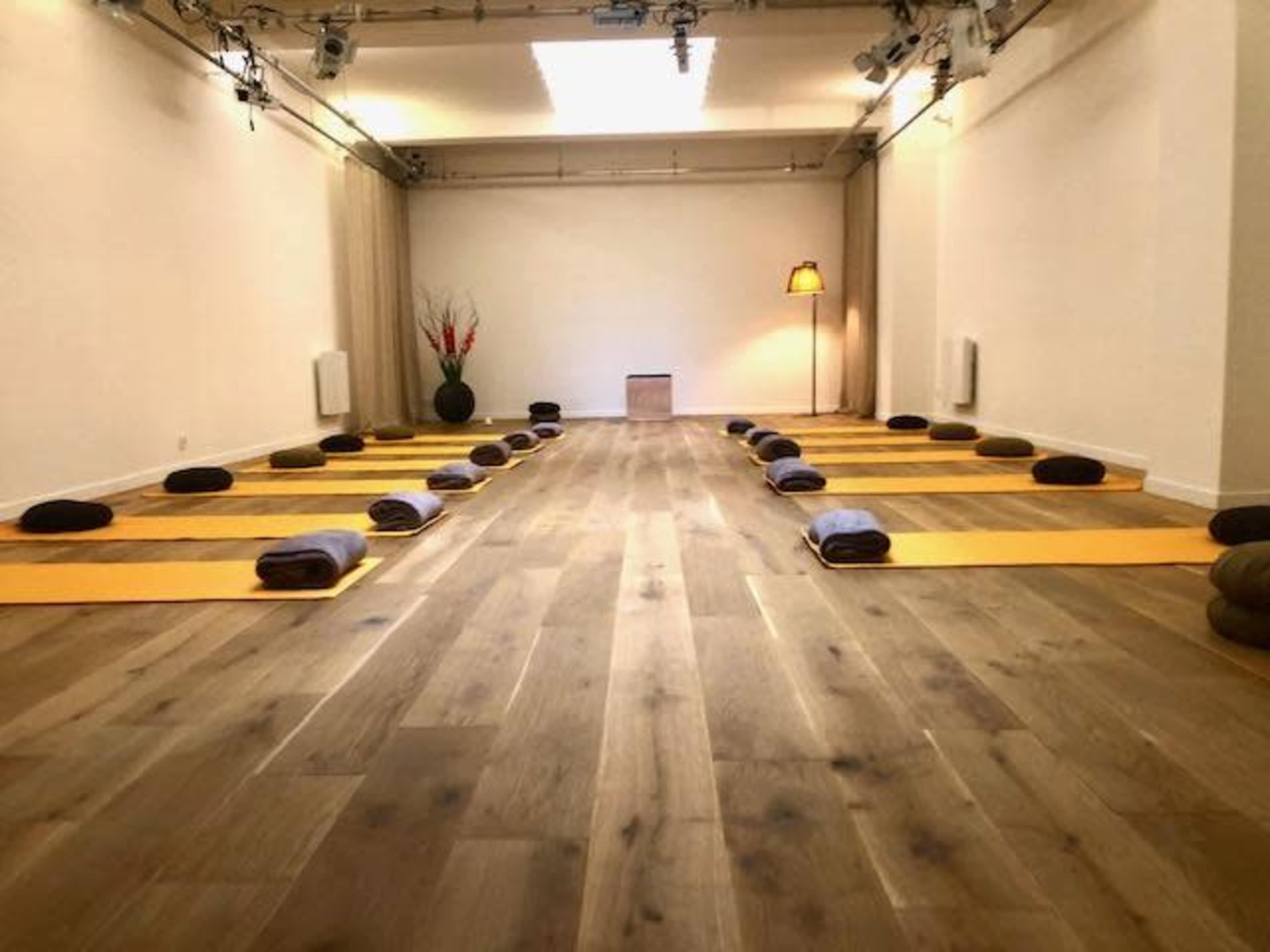 The image shows a spacious yoga studio with neatly arranged mats and props on a wooden floor, illuminated by soft overhead lighting.
