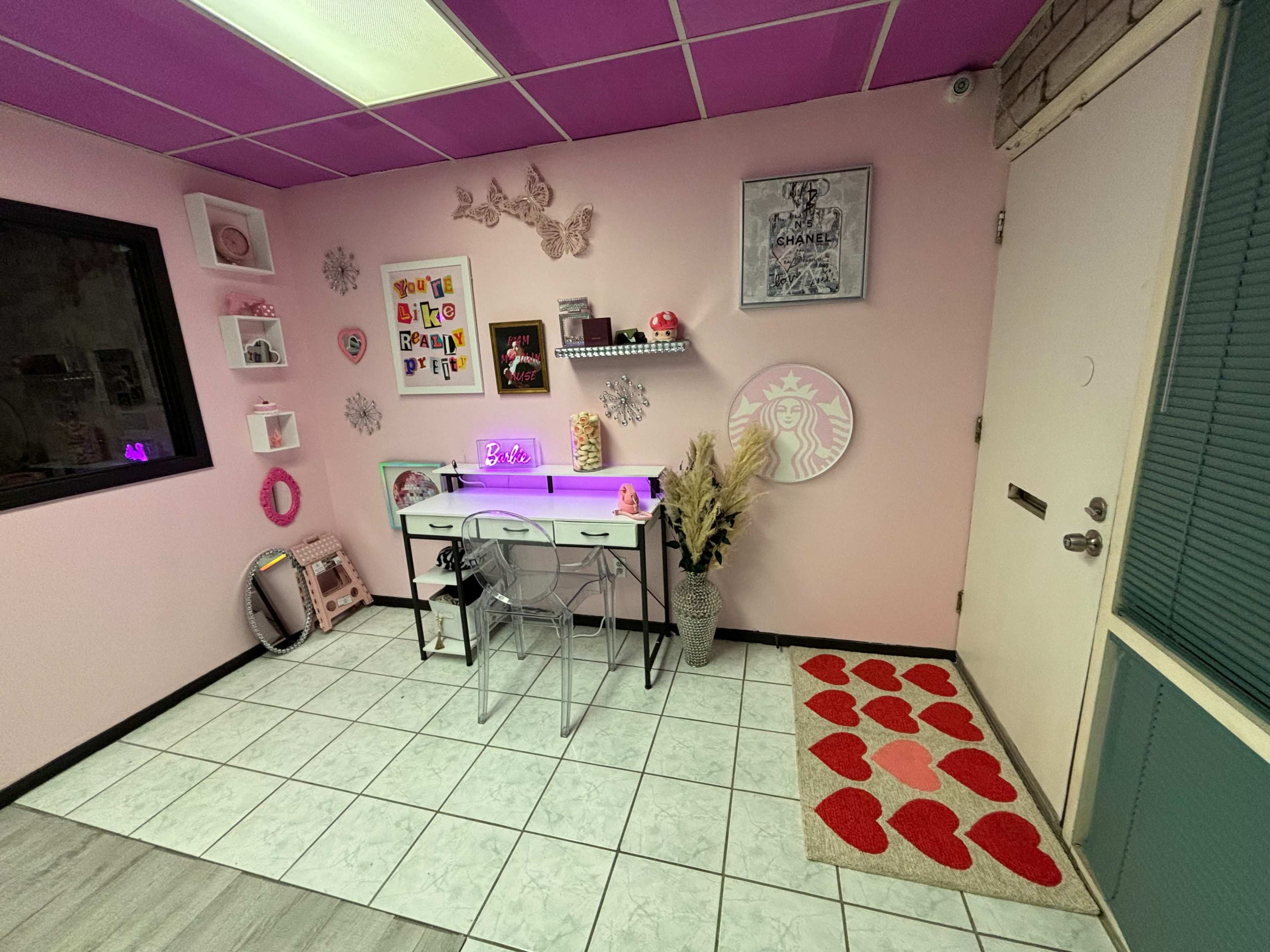 The image shows a room with pink walls, a tiled floor, a desk with a light, and decorated with various wall art and a heart-patterned rug.