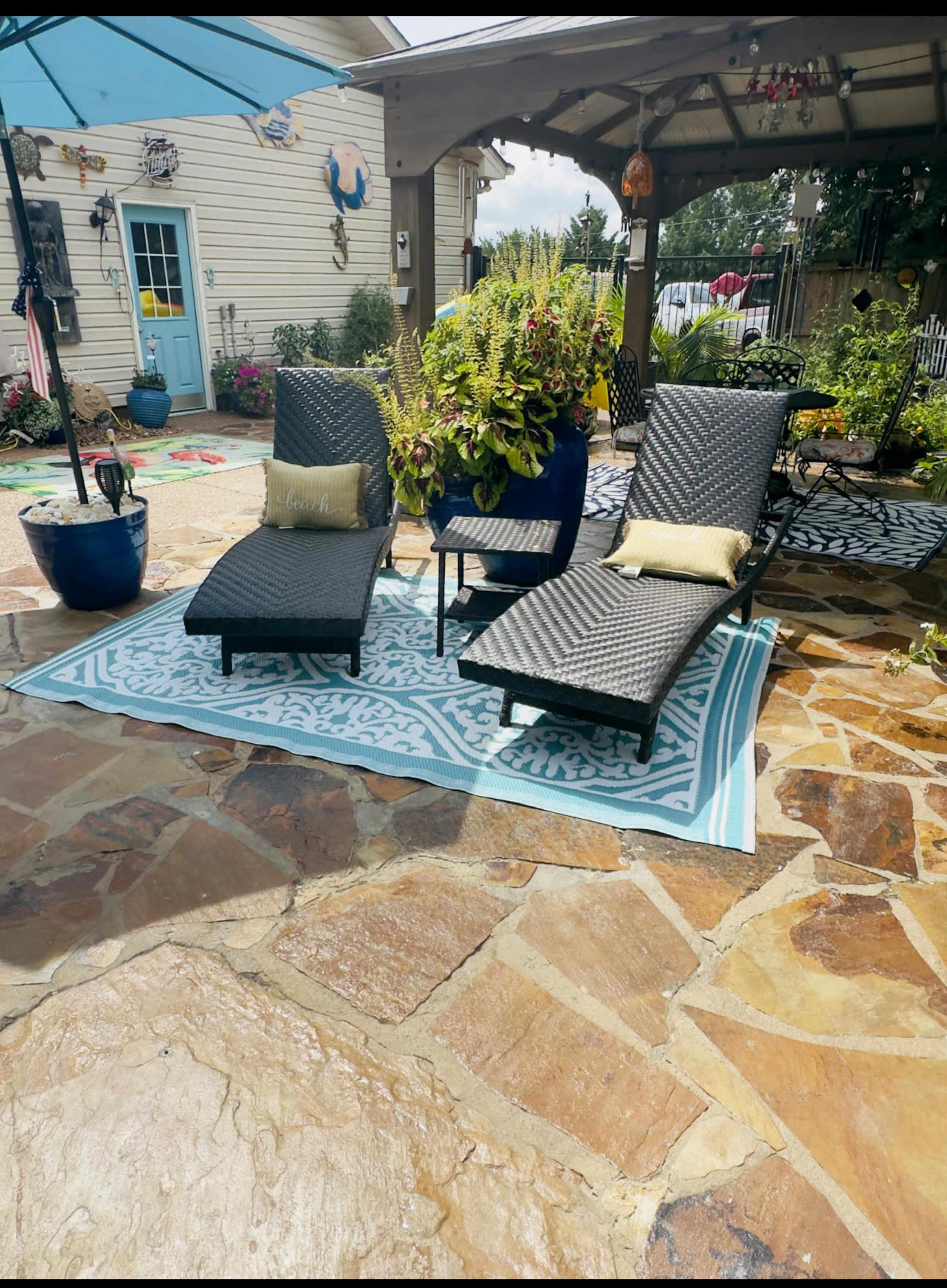 The image shows a patio area featuring three outdoor chairs on a patterned rug surrounded by potted plants and a stone floor.