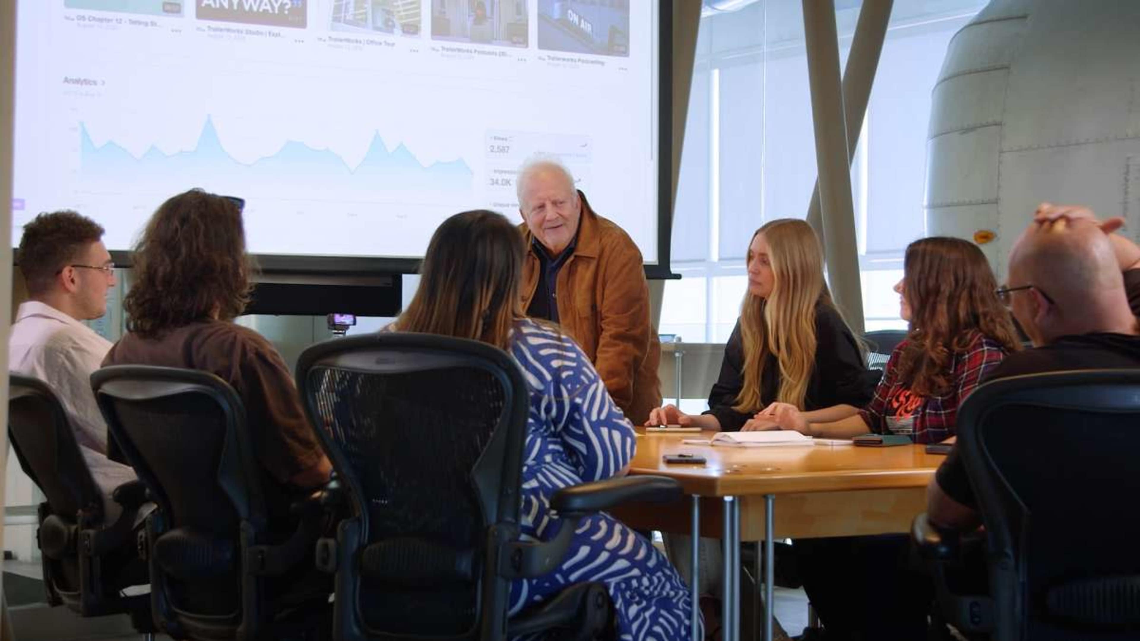 A group of seven people is seated around a conference table, discussing data displayed on a screen in a modern meeting room.