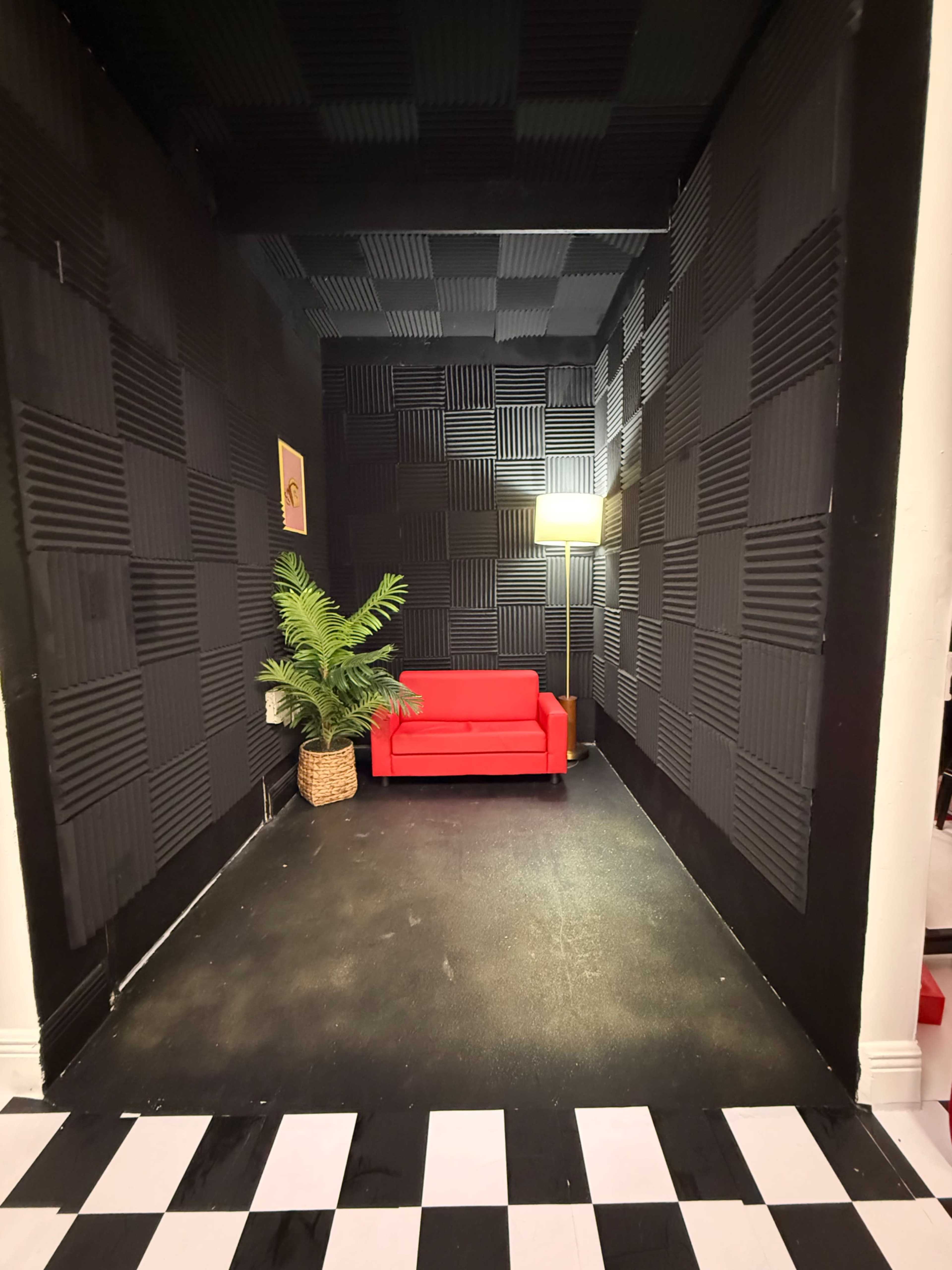 The image shows a small, black-walled room with a red couch, a potted plant in a basket, and a lamp providing light.