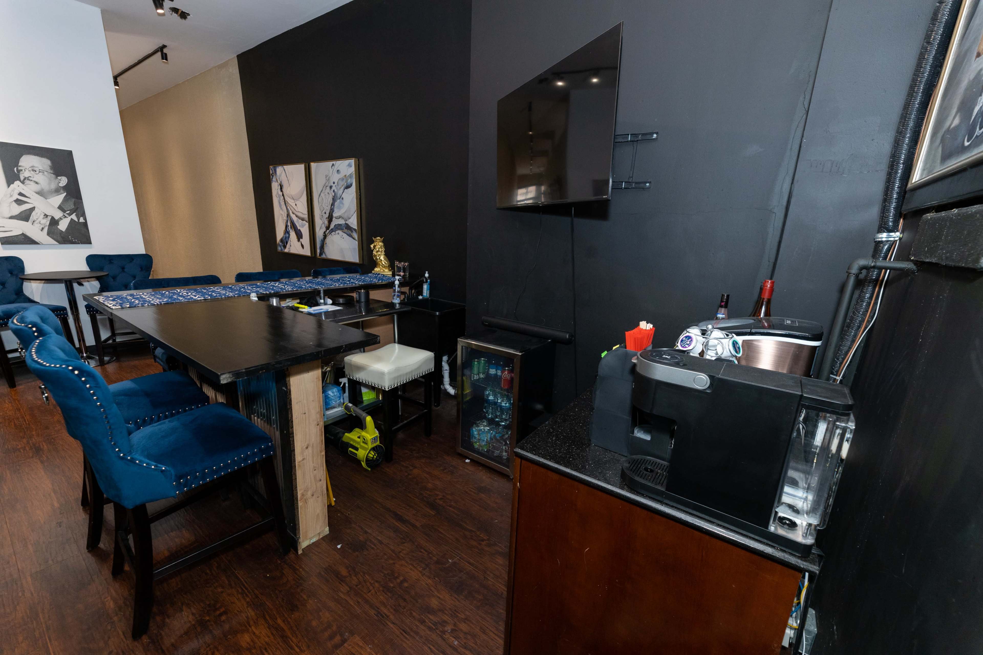 A modern interior space featuring a table with blue upholstered chairs, a wall-mounted television, and a bar area with a drink dispenser and various bottles.