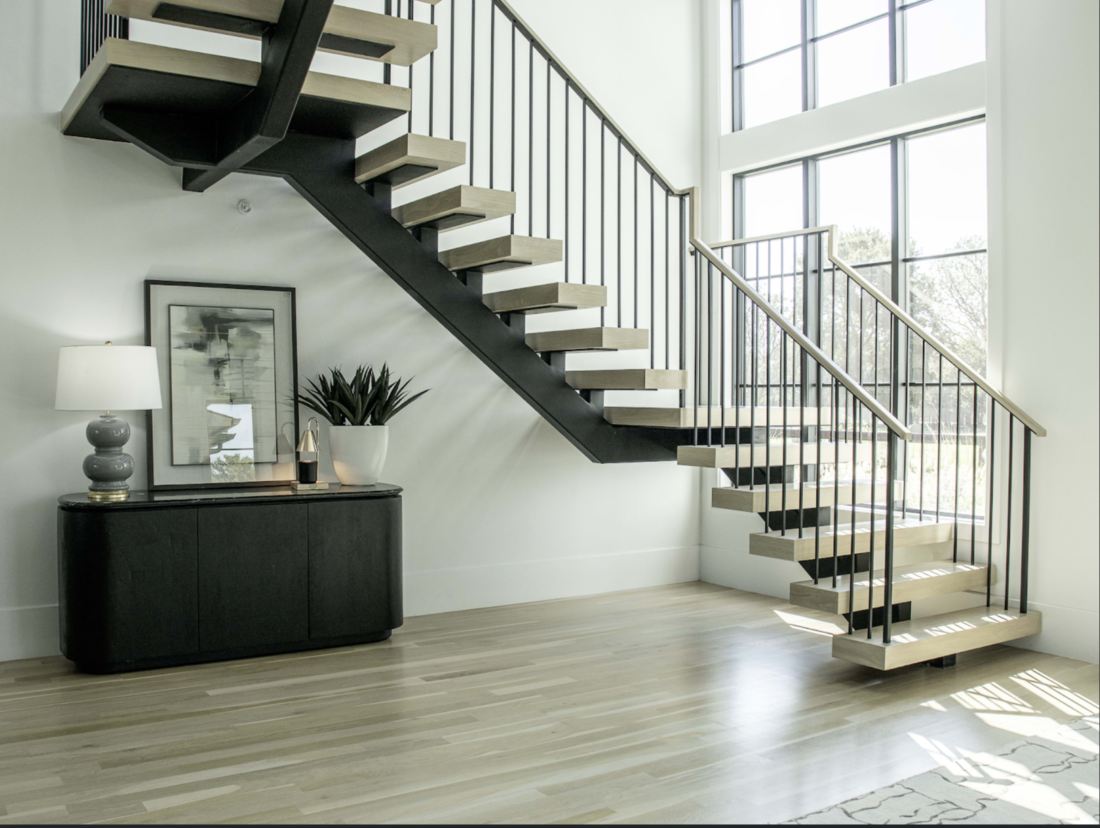 A modern staircase with wooden steps and metal railing is positioned beside a console table and large windows that allow natural light to illuminate the space.