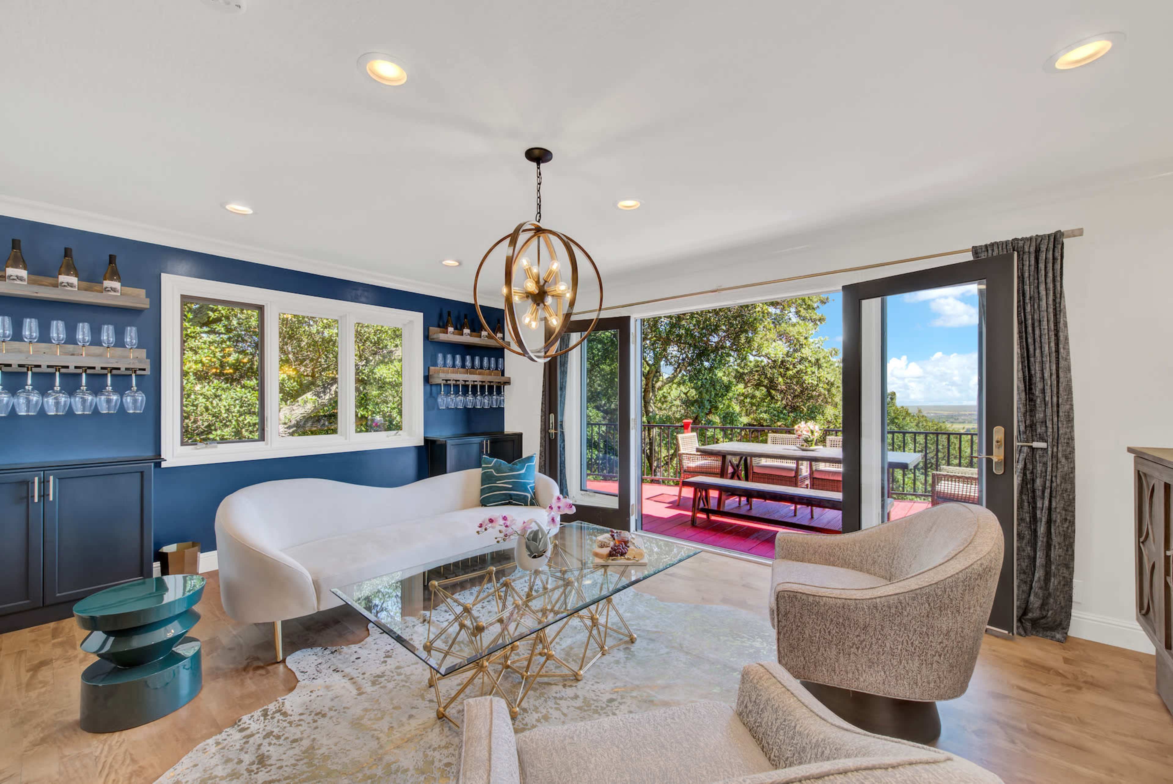 A modern living room features a glass coffee table, a white curved sofa, and large windows that open to an outdoor patio with a dining area and scenic views.