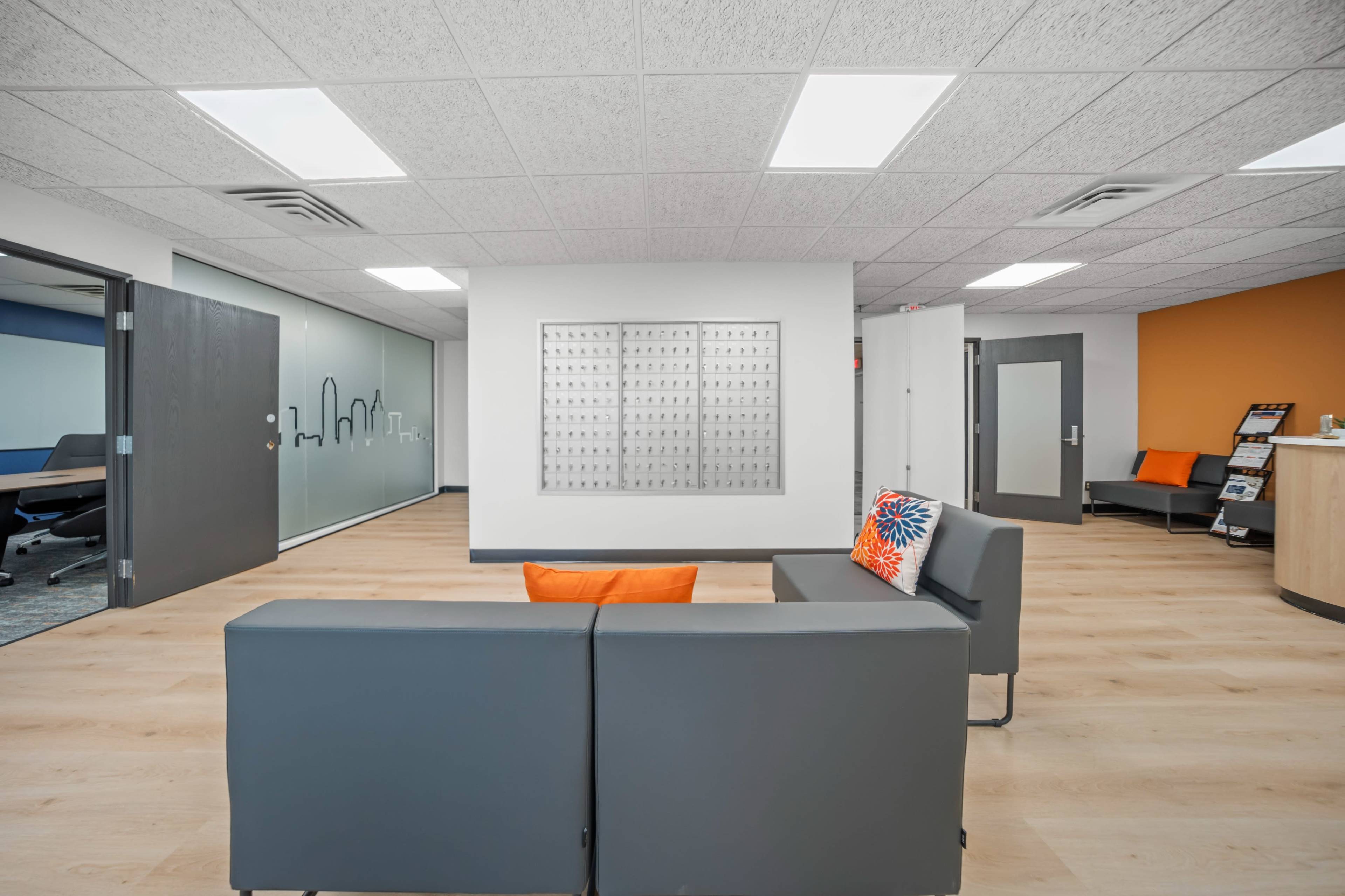 The image shows a modern office reception area with a gray couch, an orange accent wall, and a wall-mounted display panel.