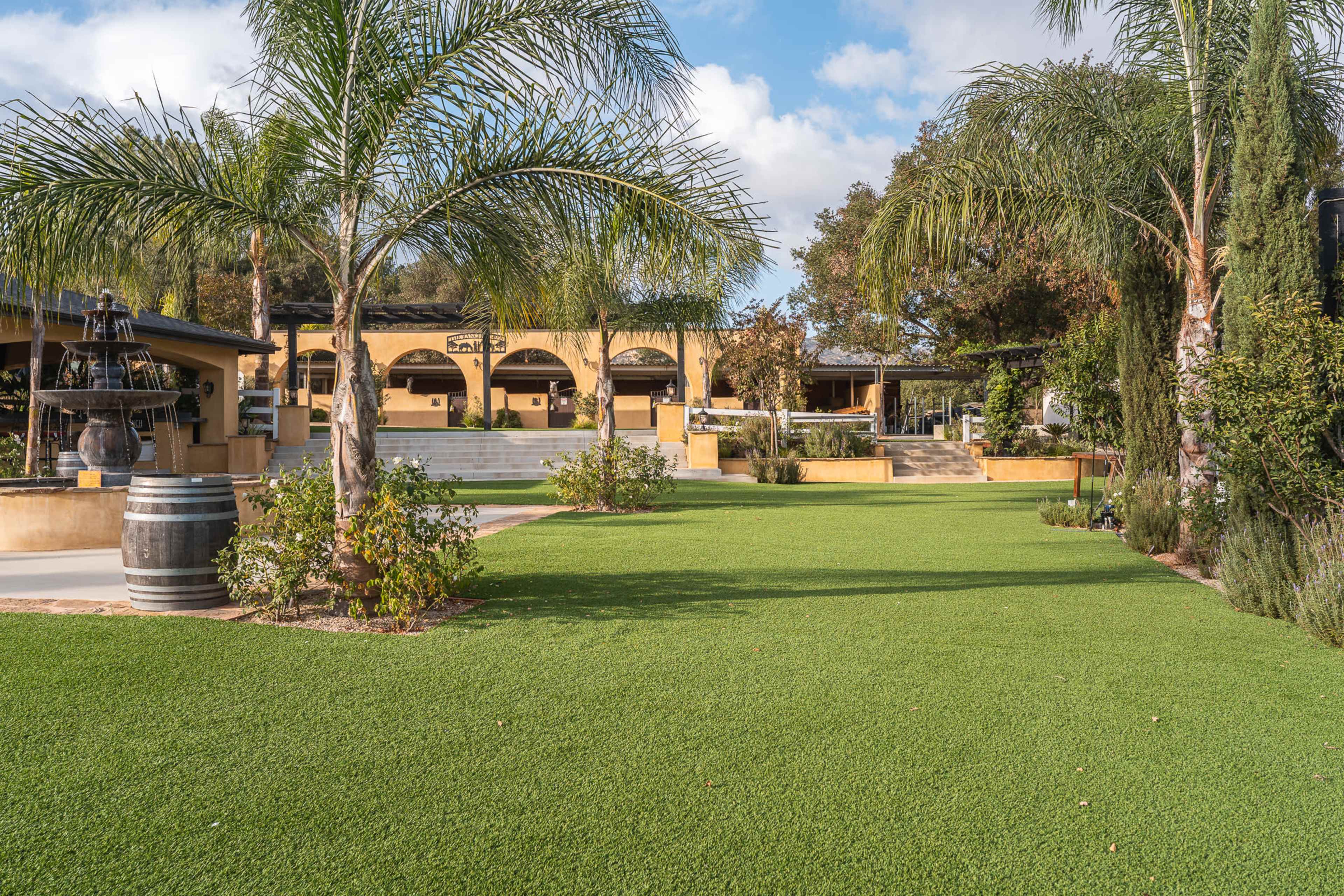 The image shows a landscaped outdoor area featuring artificial grass, palm trees, and a multi-level patio with steps leading to a building in the background.