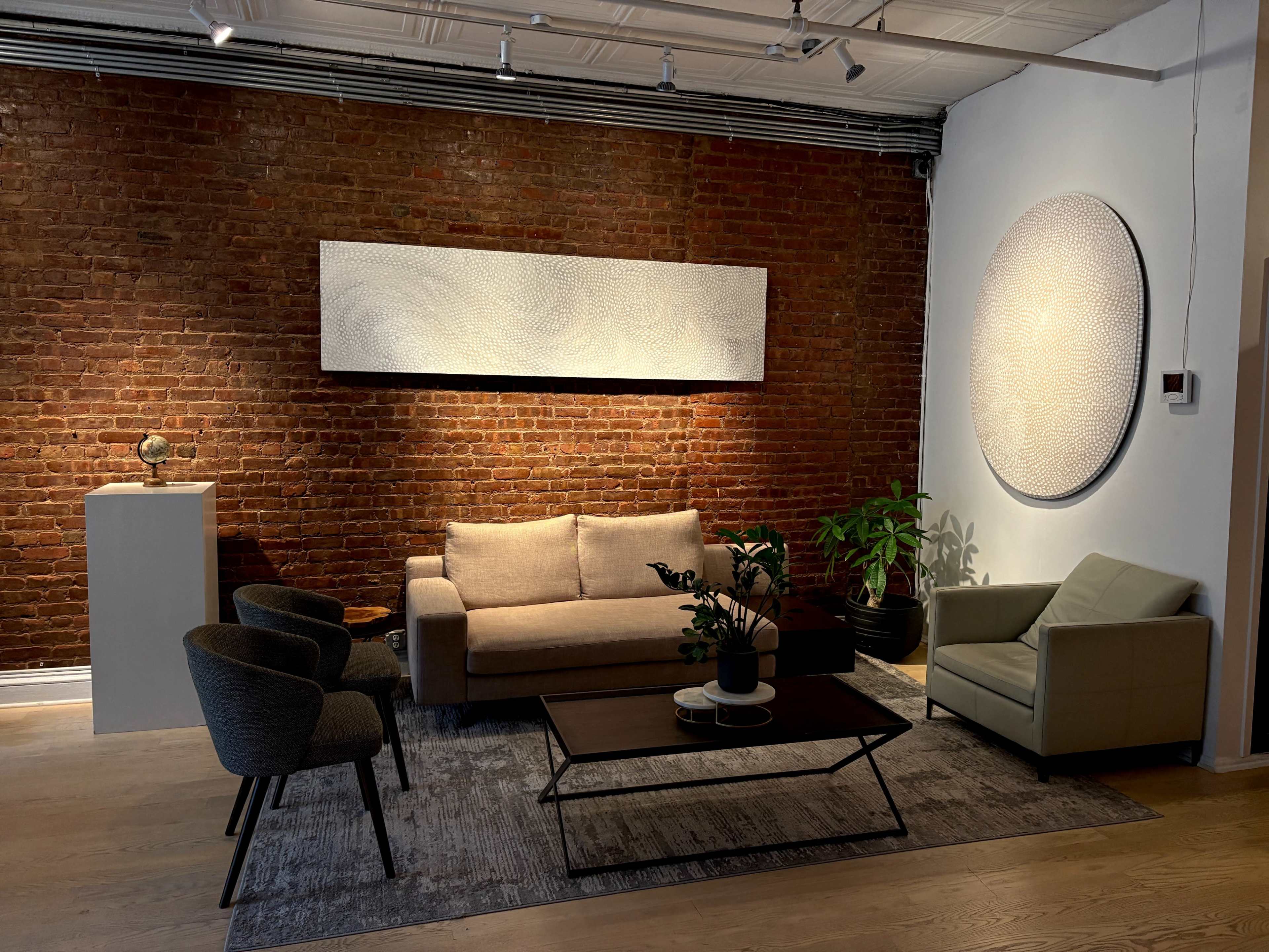 The image shows a modern living space featuring a beige sofa, two dark armchairs, a coffee table, and two round wall artworks against a backdrop of exposed brick.