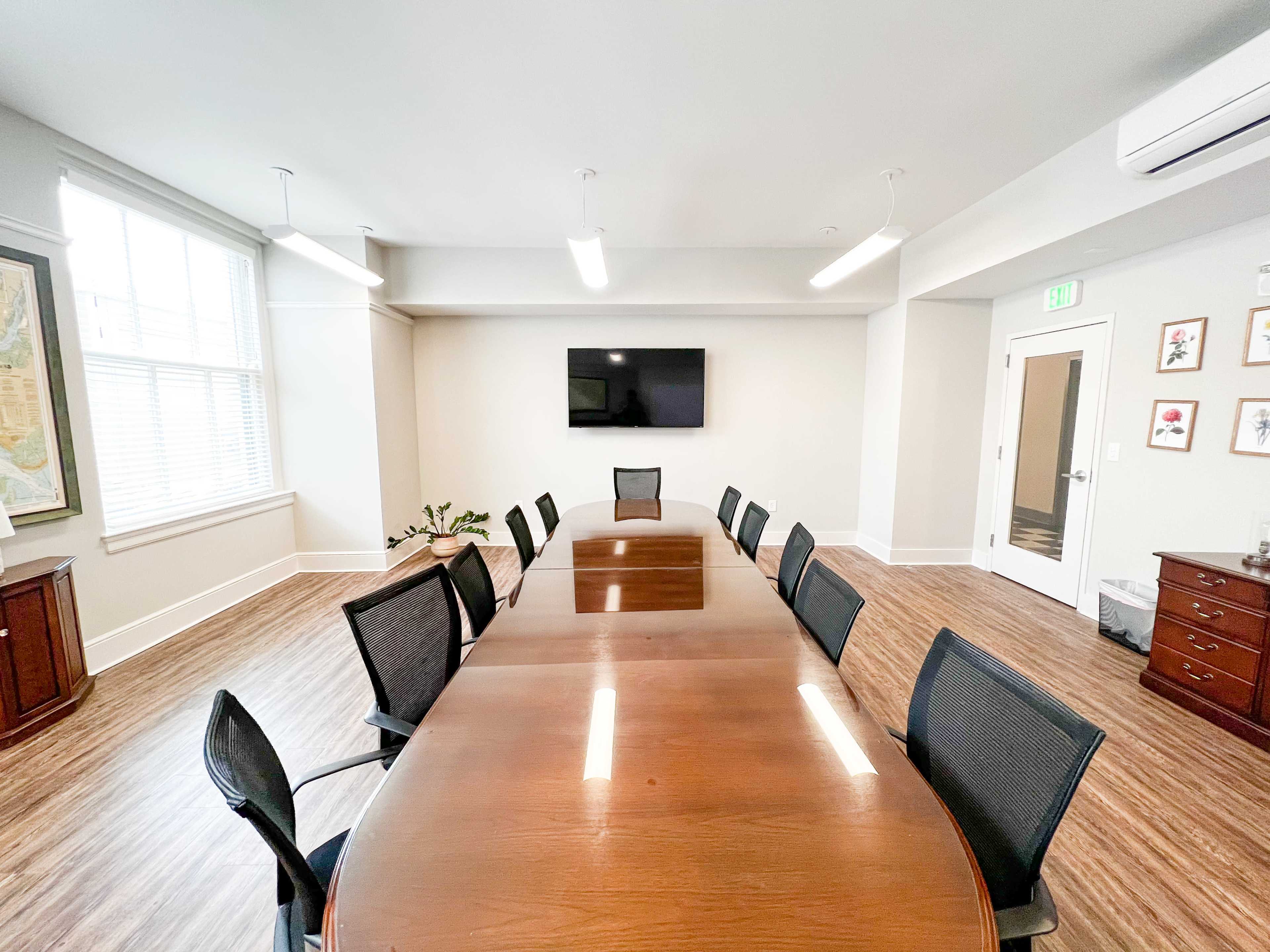 A conference room featuring a long wooden table surrounded by black chairs, with a wall-mounted television and windows letting in natural light.
