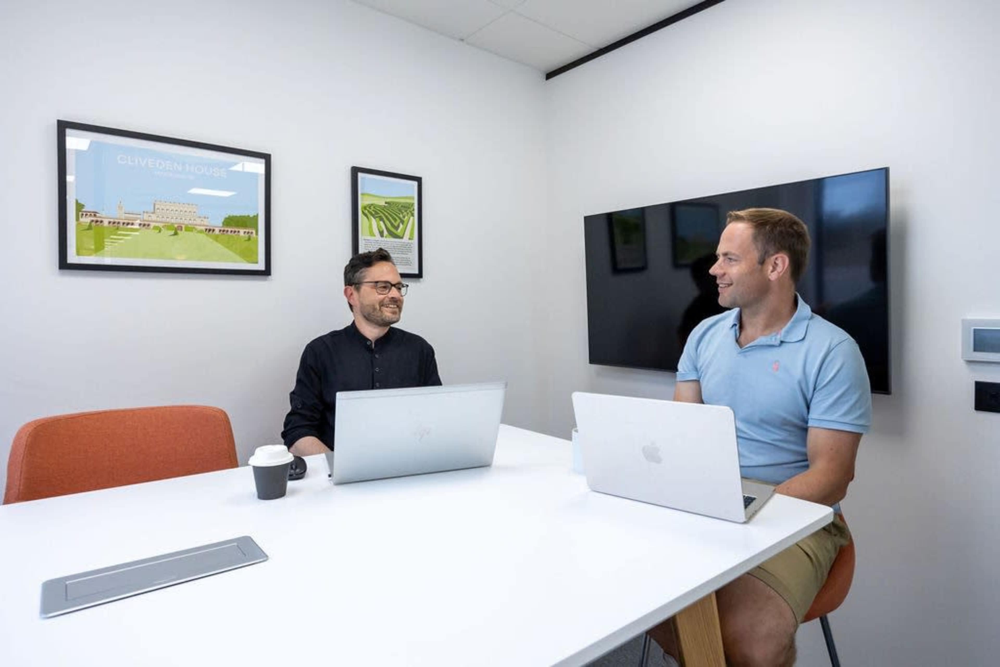 Two men are sitting at a white table in a bright office space, each working on a laptop while engaging in conversation.