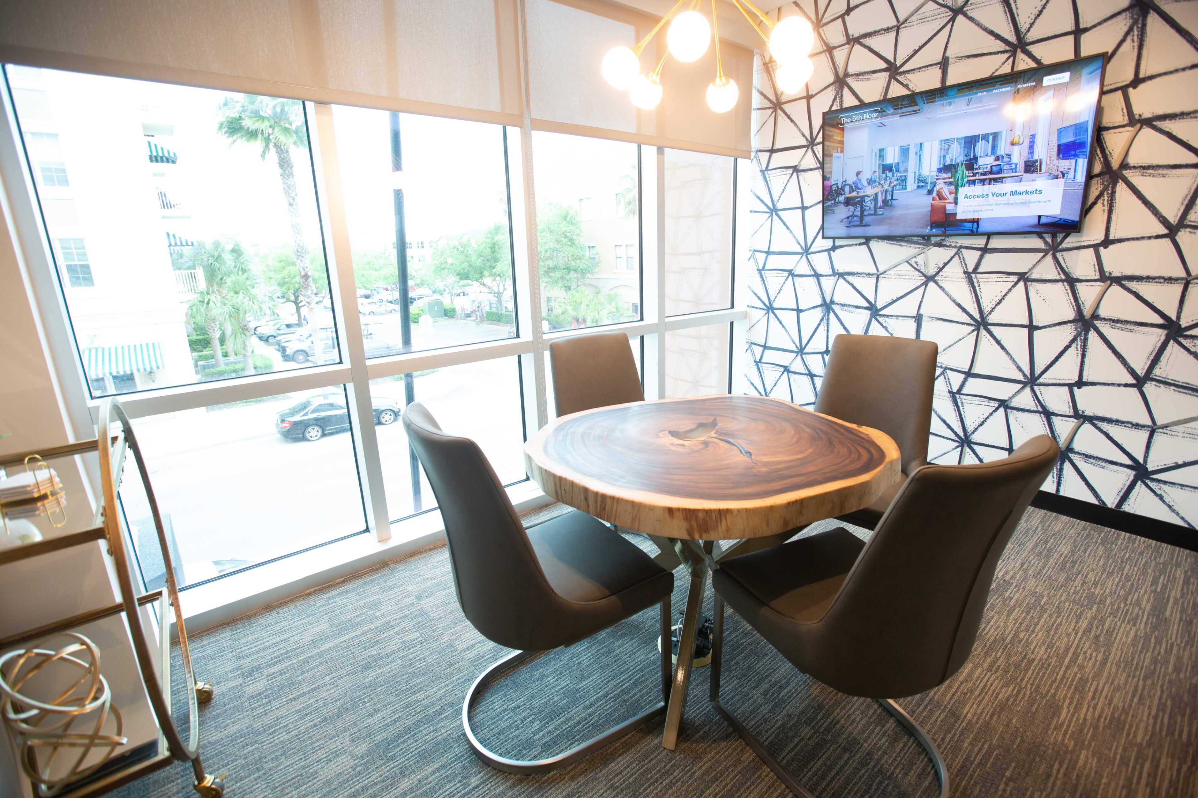 A modern meeting area features a round wooden table surrounded by four black chairs, positioned next to large windows displaying an outdoor view.