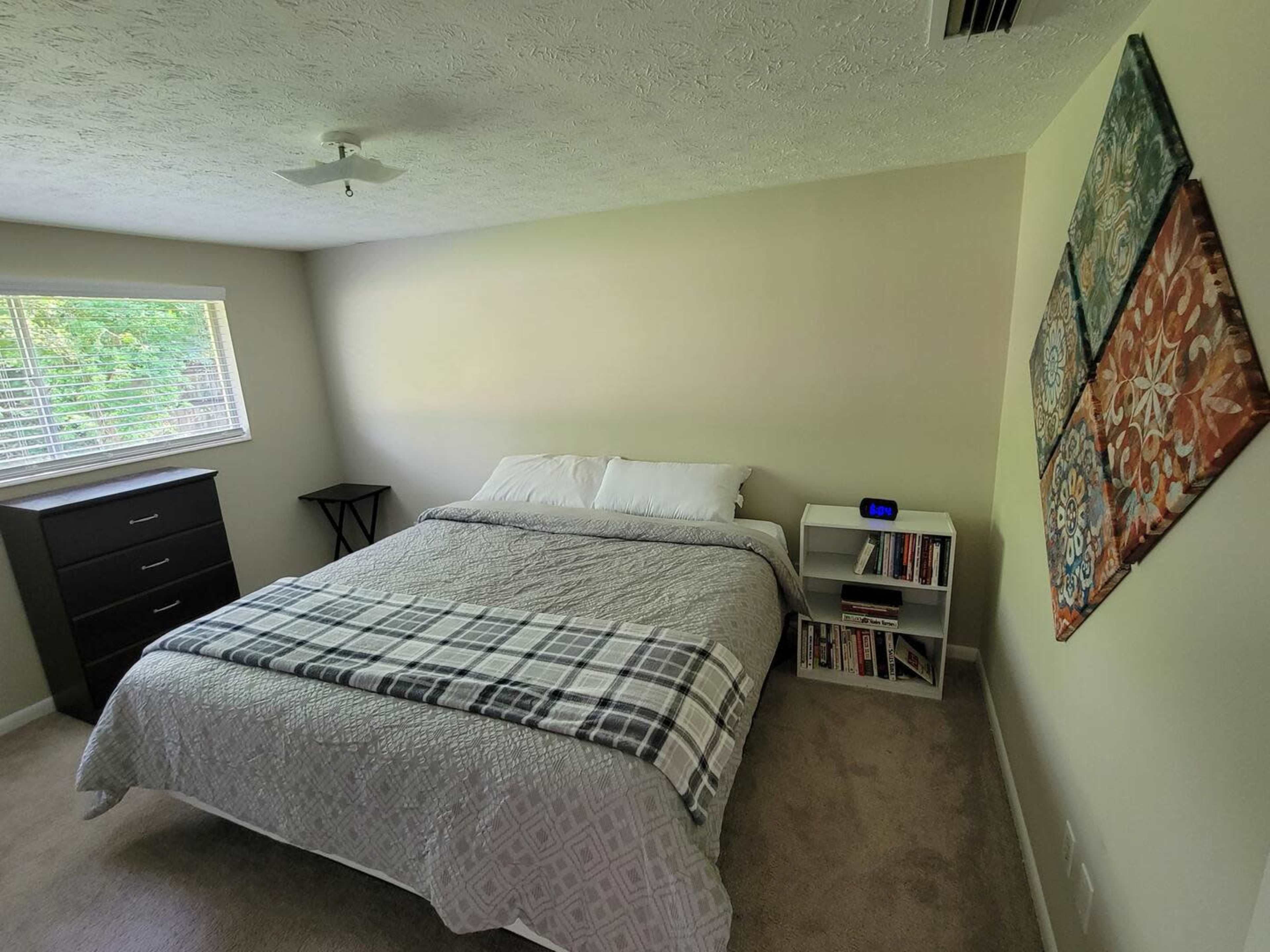 A simple bedroom featuring a bed with a gray quilt, a nightstand with a clock, a white bookshelf, and a dresser, with a window providing natural light.