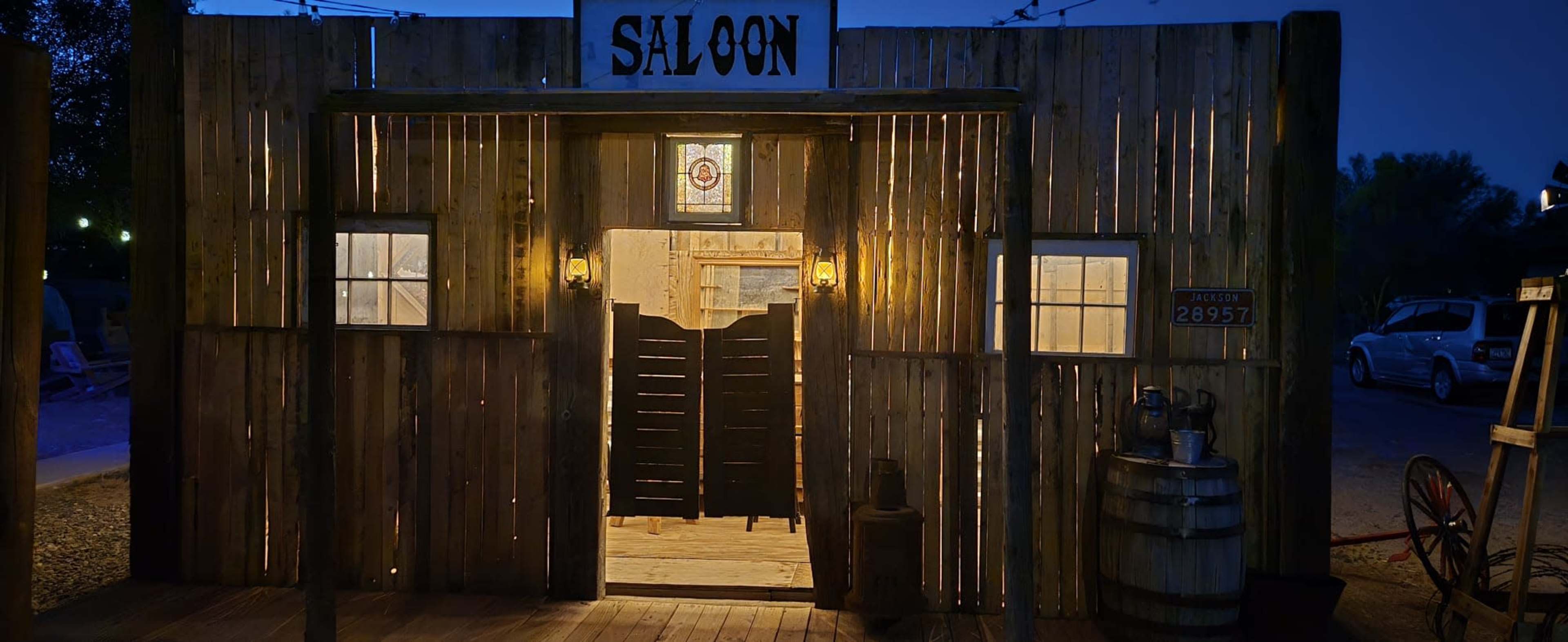 A wooden saloon with a sign above the entrance is illuminated by lights at dusk, showcasing a rustic facade and an open door with a half door inside.