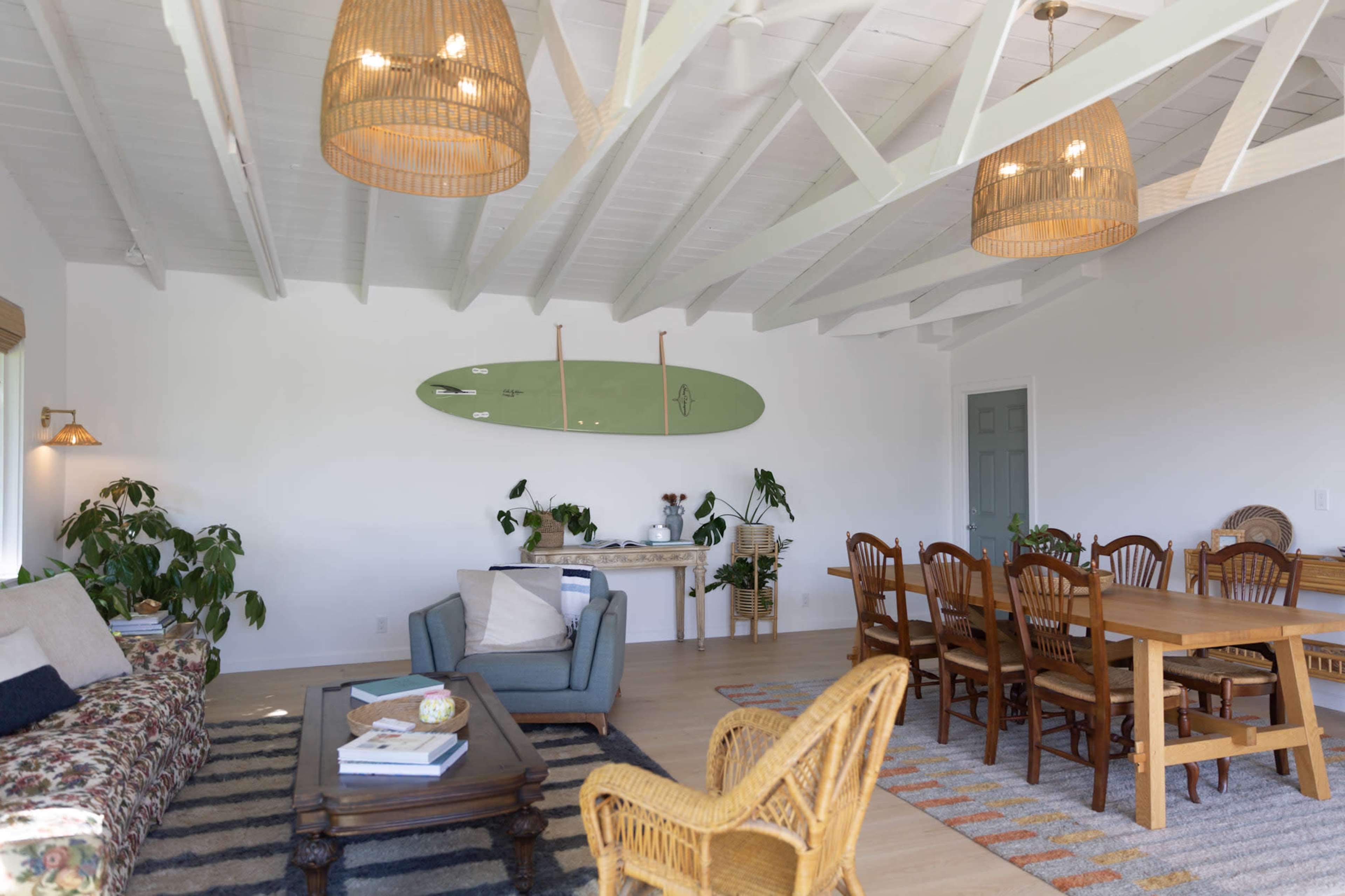 The image shows a bright, open living space with a dining area, wooden furniture, a surfboard mounted on the wall, and plants scattered throughout.