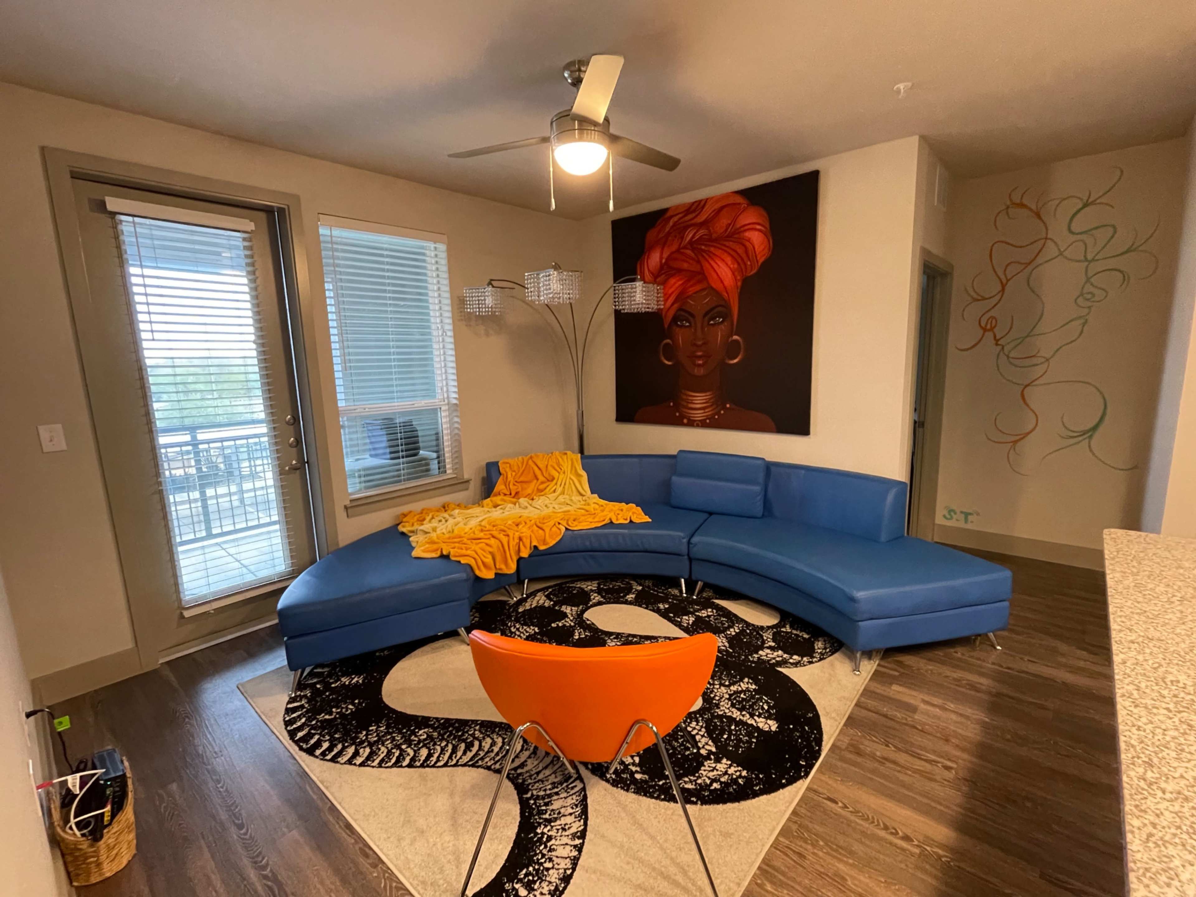 The scene shows a modern living room featuring a blue sectional sofa, an orange chair, a large painting of a woman on the wall, and a patterned rug on a wooden floor.