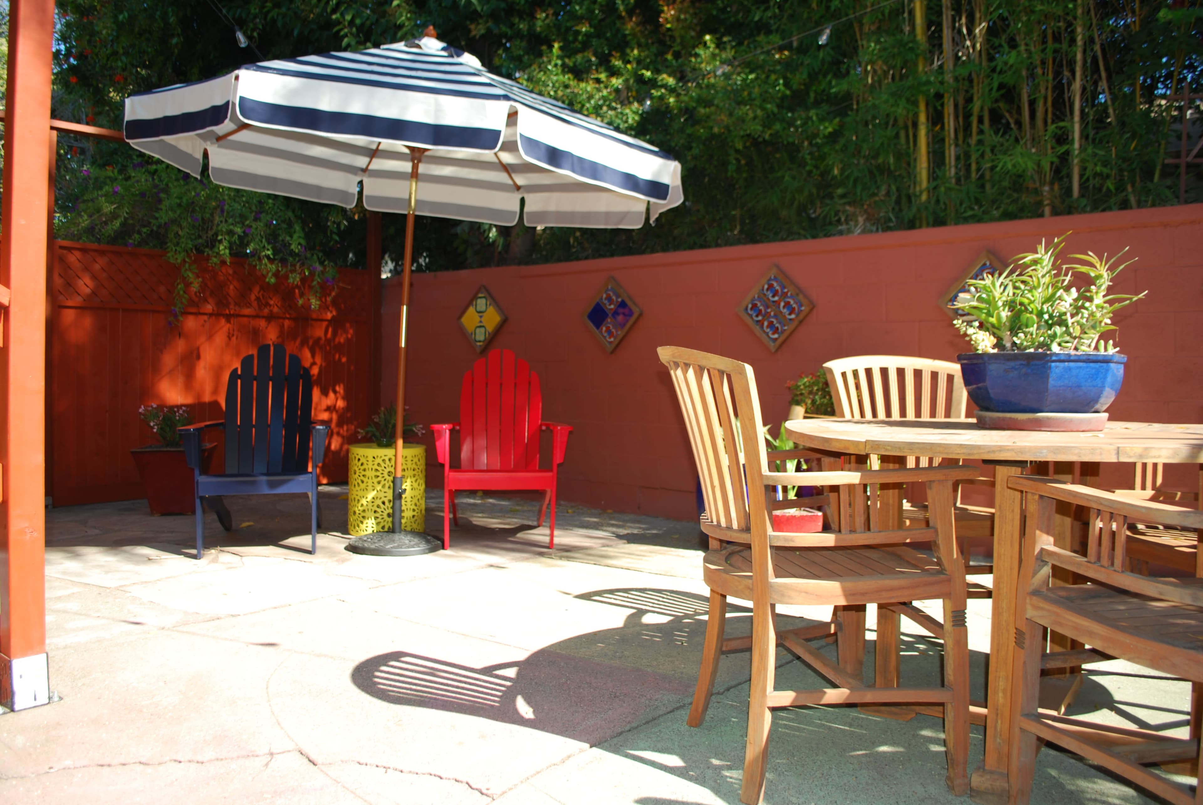A small outdoor patio features a round wooden table surrounded by chairs, with two colorful adirondack chairs and a large striped umbrella in a corner.