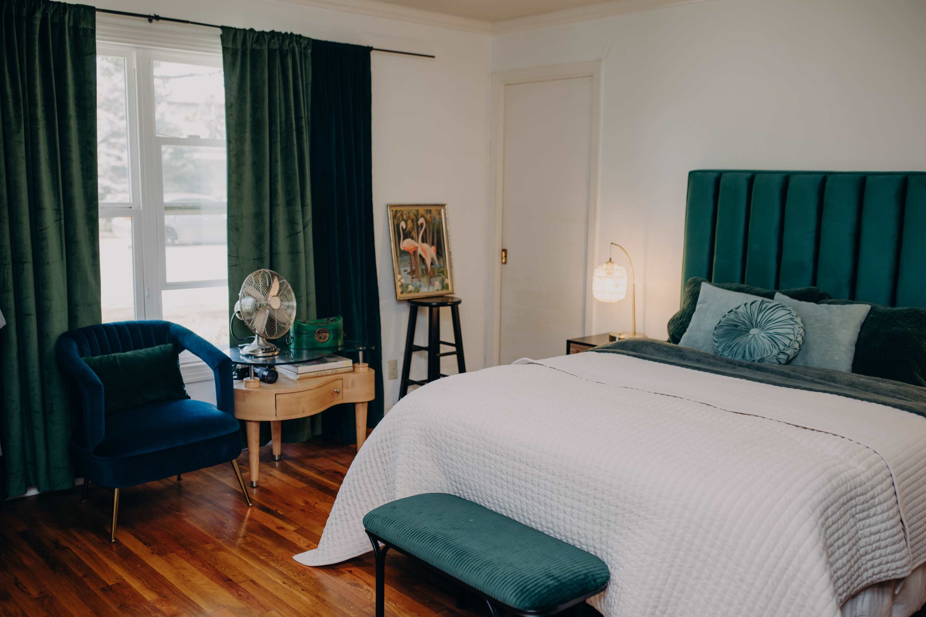 A cozy bedroom features a large green headboard, a neatly made bed, a blue accent chair, and a wooden nightstand beside a window.