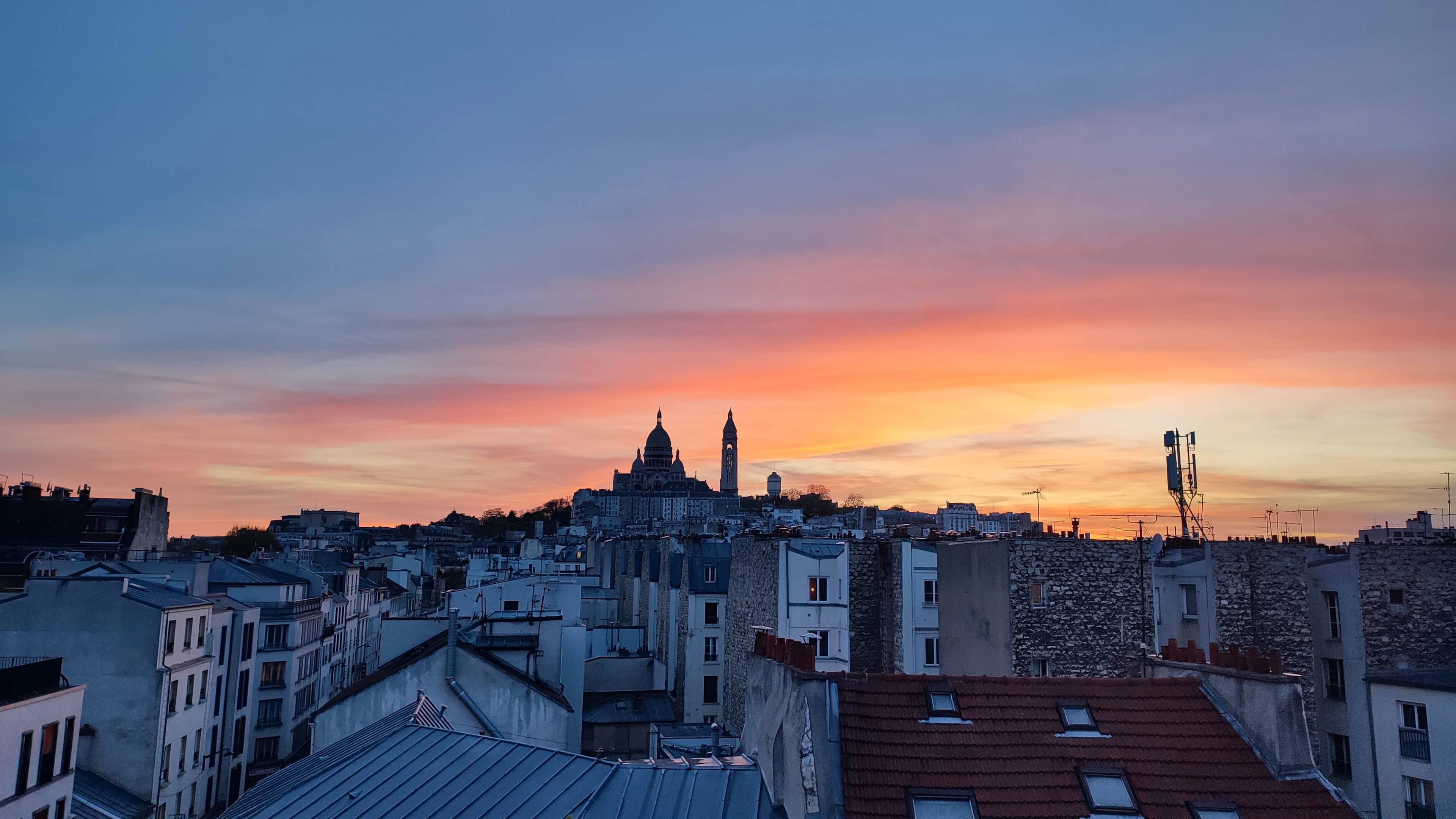A panoramic view of a city skyline at sunset with rooftops in the foreground and a silhouette of buildings, including a prominent basilica, against a colorful sky.