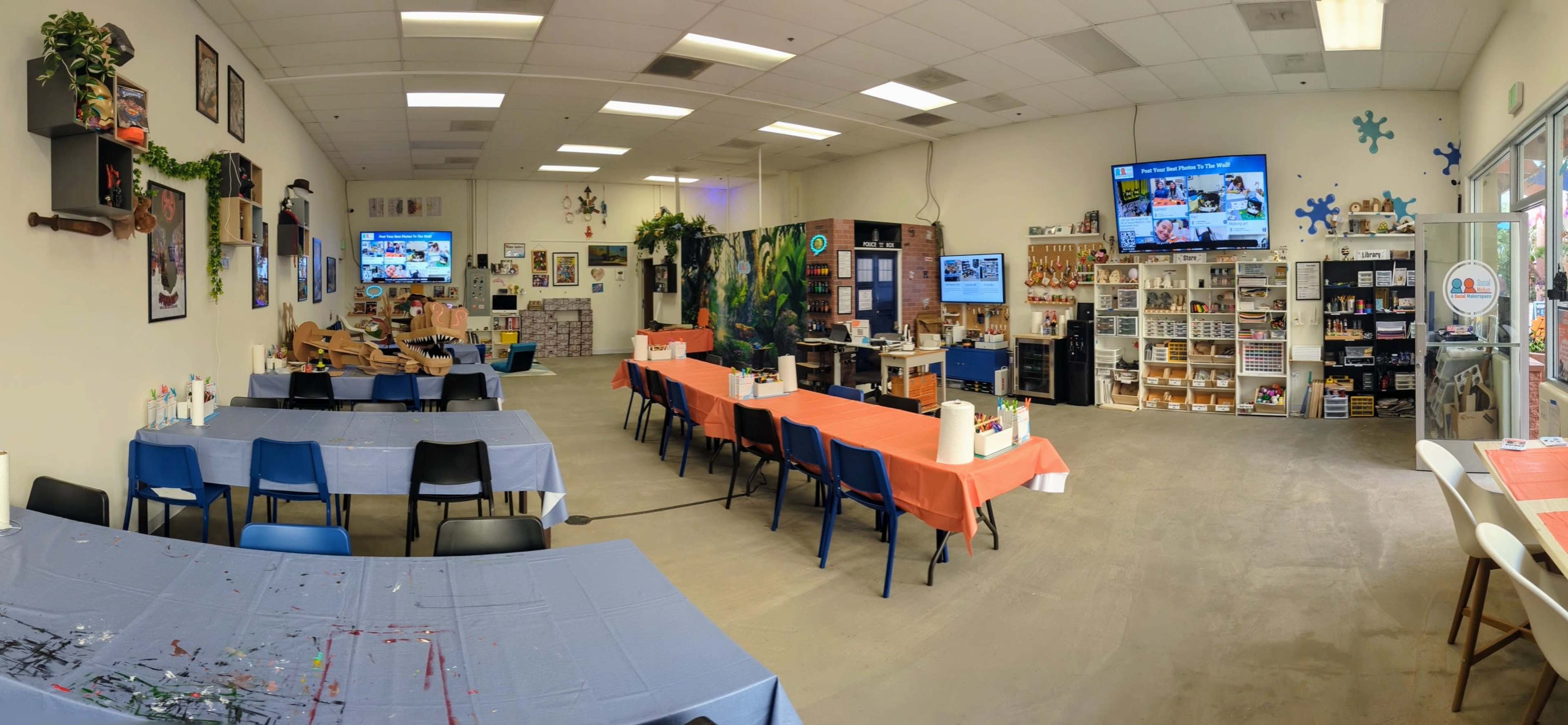 A spacious art studio with multiple tables set for painting activities, two large screens displaying content, and various art supplies organized on shelves.